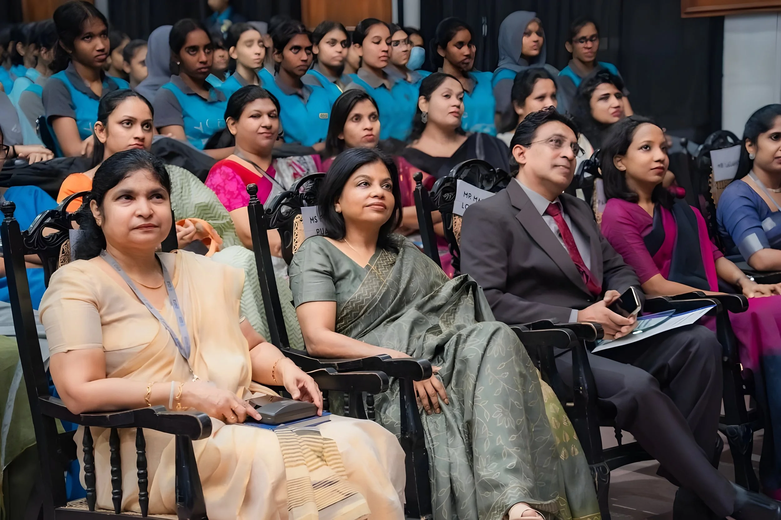 An audience of men and women sitting in a conference or seminar, some taking notes, in an indoor auditorium.