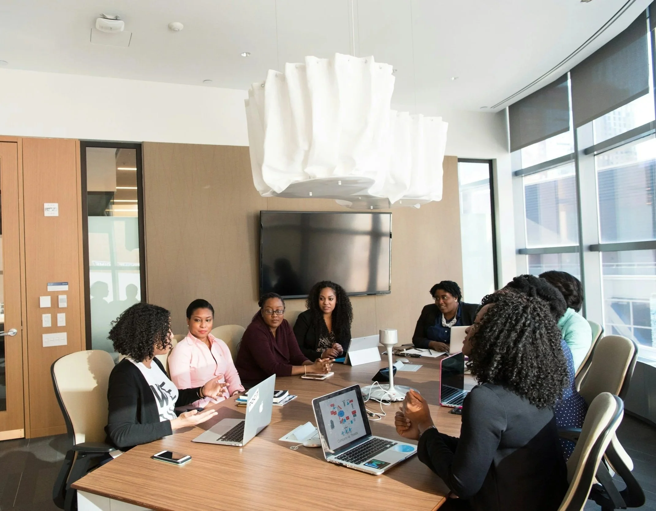 A diverse group of women in a modern conference room having a meeting, with laptops and tablets on the table. Large windows let in natural light.