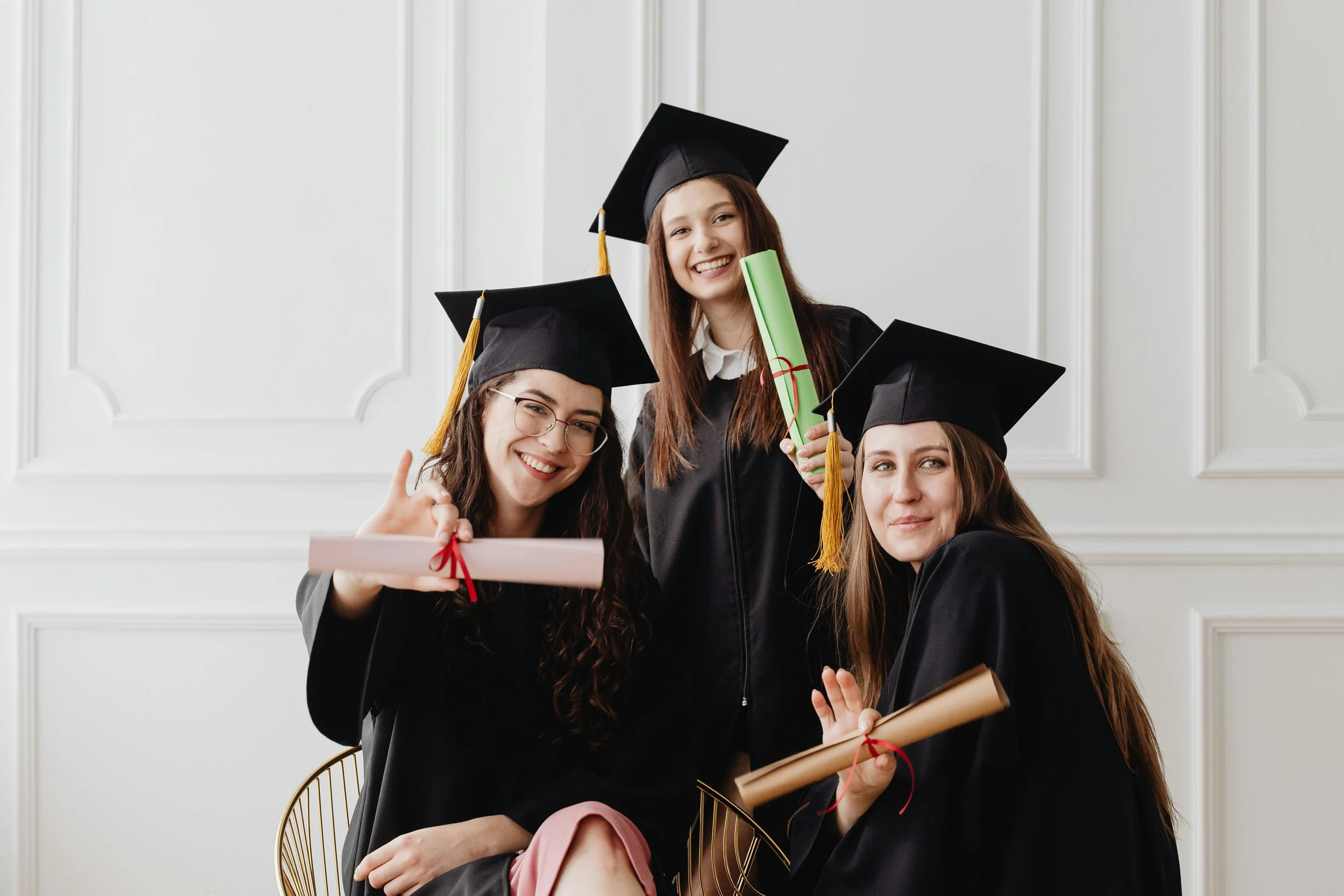 Three young women in black graduation caps and gowns celebrating graduation, holding diplomas and a rolled certificate, smiling at the camera.