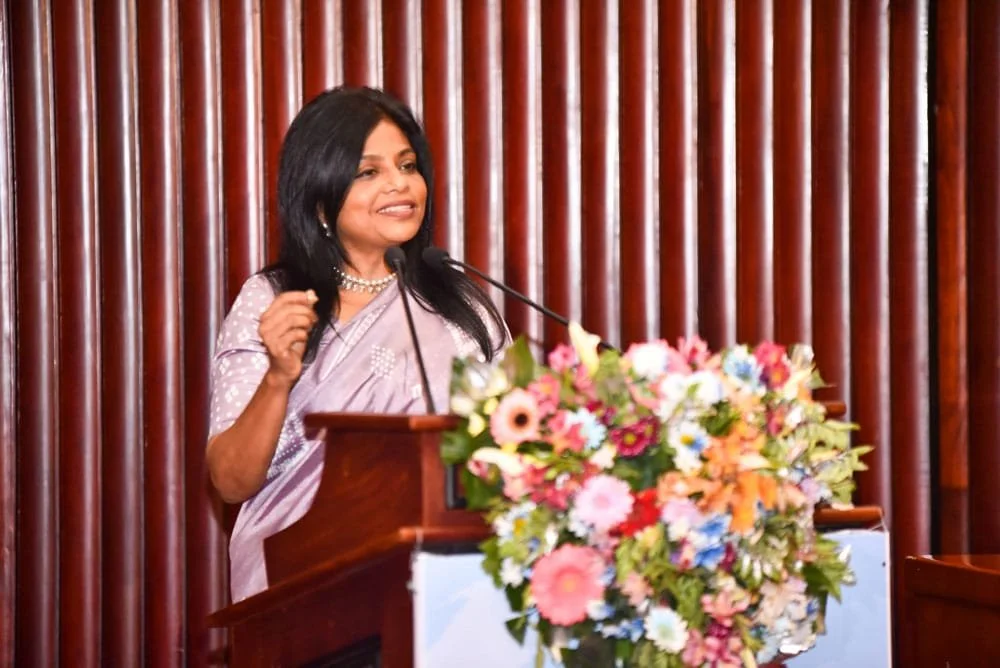 A woman speaking at a podium with a microphone, decorated with a large bouquet of flowers, in front of a wooden-paneled wall.