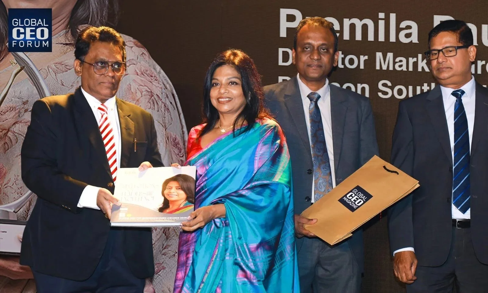 Four professionally dressed people standing on stage at the Global CEO Forum, exchanging awards and documents. There is a man in a suit with a striped tie handing an award to a woman in a colorful saree, another man holding a folder, and a third man in a suit standing nearby.