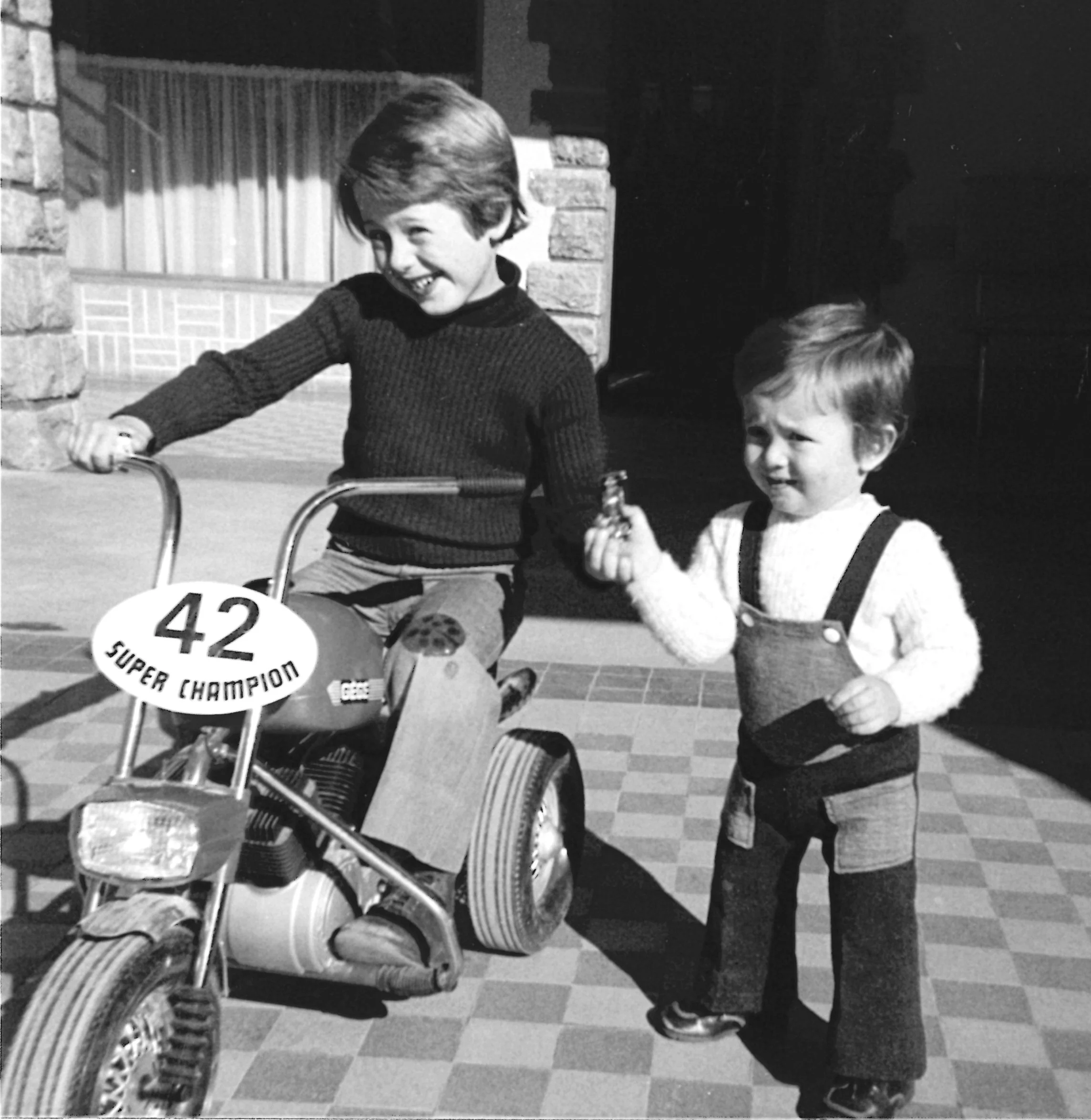 Deux jeunes garçons, un plus âgé conduisant une moto avec le numéro 42 et la mention "Super Champion", et un plus jeune tenant une petite figurine ou un jouet, debout sur une surface pavée devant une maison en pierre.