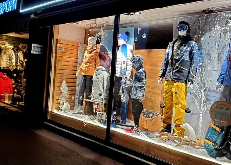 Vitrine d'un magasin de vêtements d'hiver avec des mannequins portant des vestes, pantalons de ski, et accessoires de ski. Décor hivernal avec des éléments de neige et des arbres.