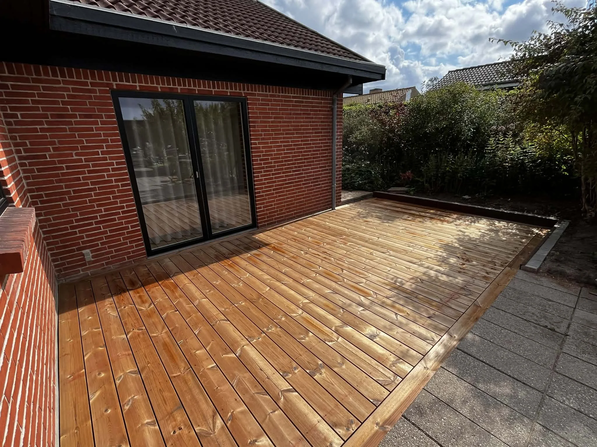 Et nyt trædækket terrasse foran et rødt murstenshus med glasdør, omgivet af buske og planter under en skyet himmel.