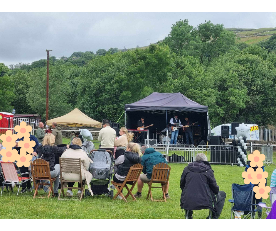 An outdoor concert in a park with a small stage where a band is performing. Audience members watching the performance are seated on lawn chairs and benches on a grassy area. Some people are standing near the stage, and a few are taking photos. The background features green trees and hills under an overcast sky.