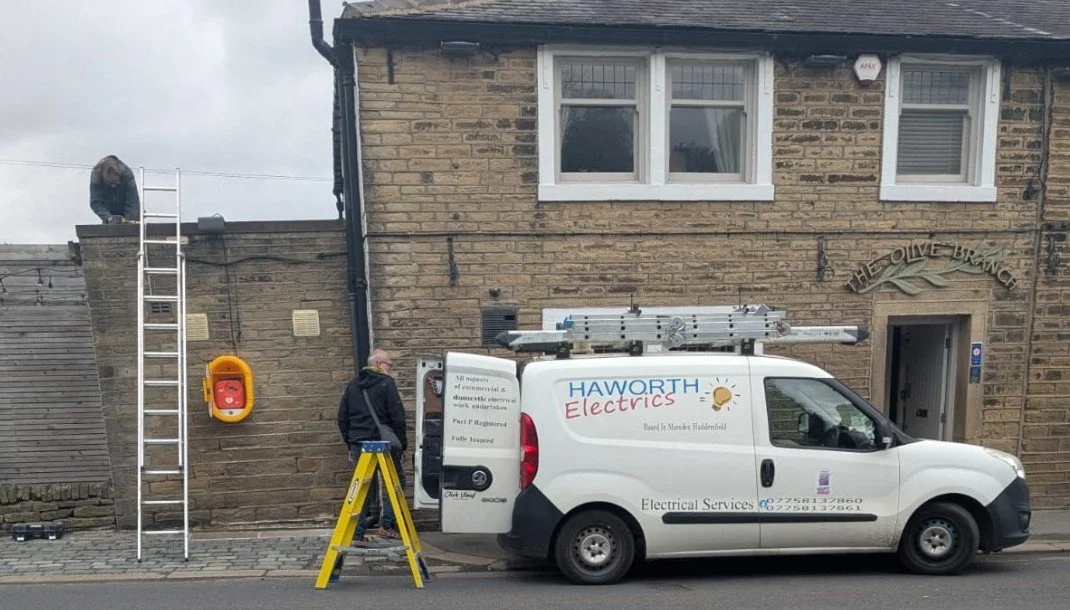 Electricians working outside 'The Olive Branch' in Marsden, with ladders and a white van marked 'Haworth Electrics' parked on the street.