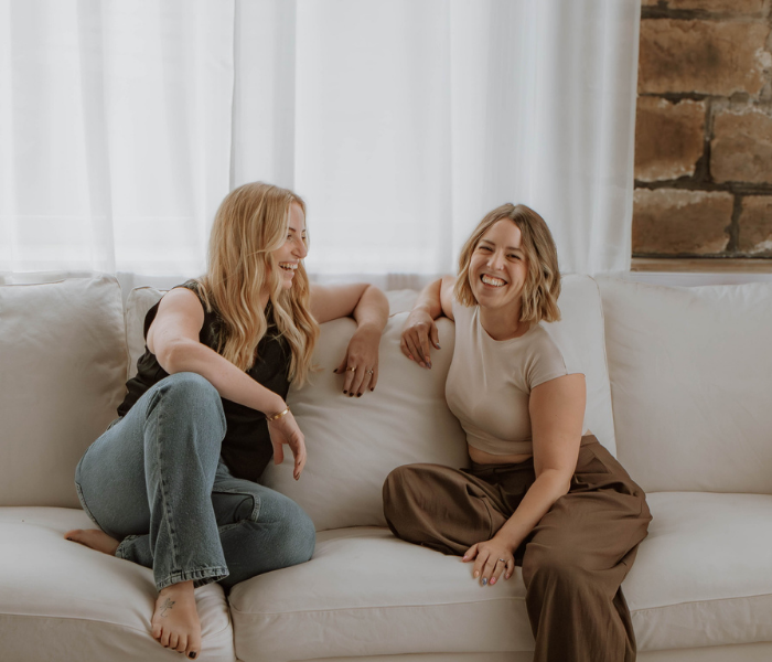 Two women smiling together, sat on a sofa. They own Not Another Social; a boutique creative marketing agency in Huddersfield, West Yorkshire