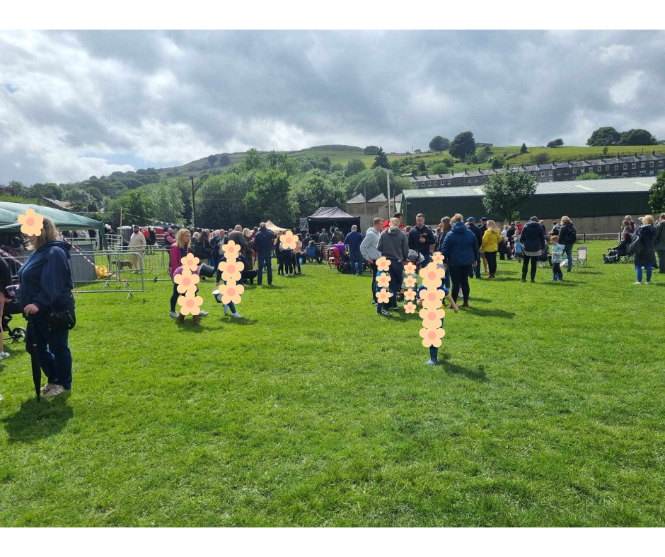 People gathered outdoors in Marsden on a grassy field at a community event, with tents and stalls visible, and hills in the background under cloudy skies.