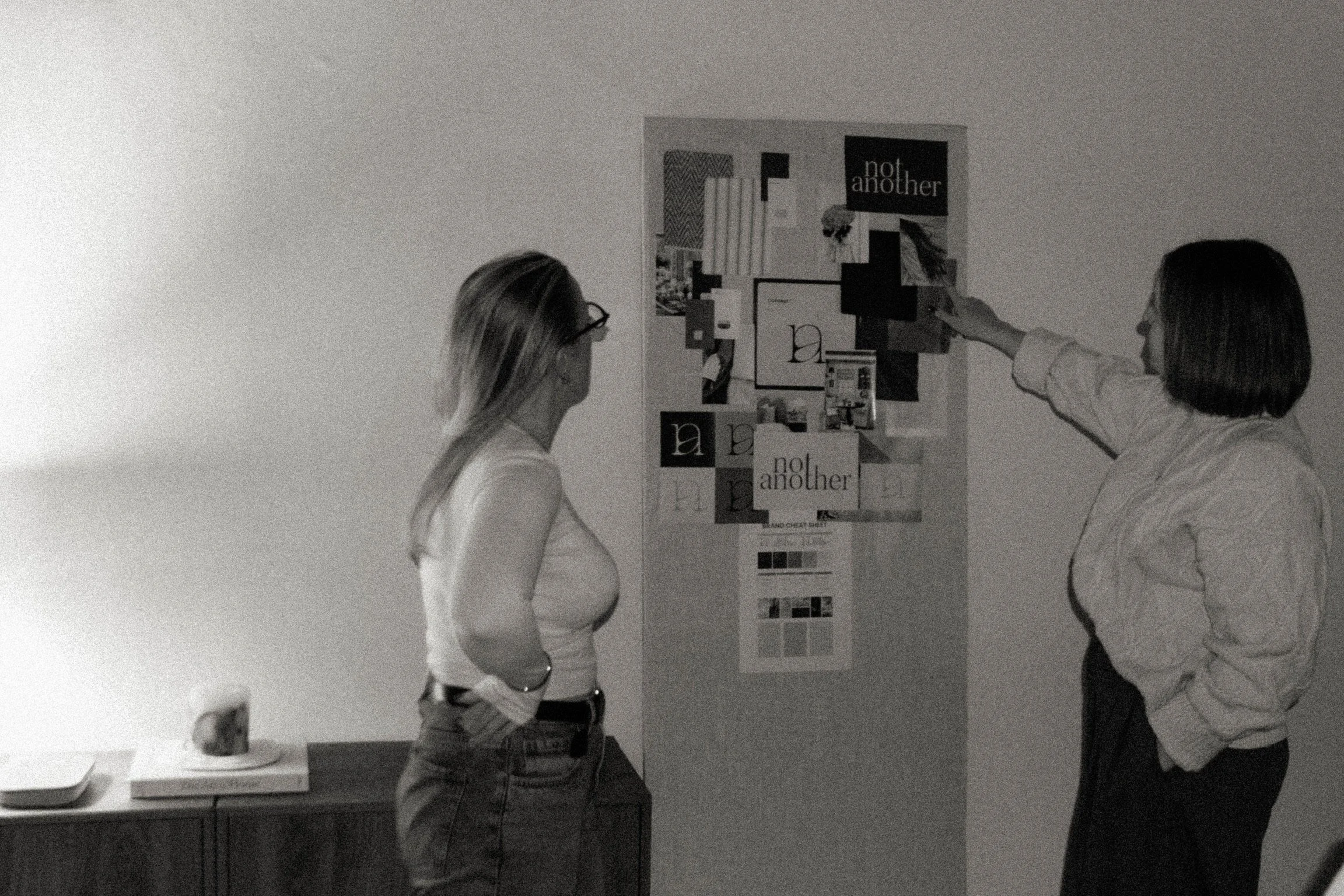 The two owners of Not Another Social in Huddersfield, West Yorkshire looking at a large pin board with images on it. The image is in black and white and has been edited to be grainy.