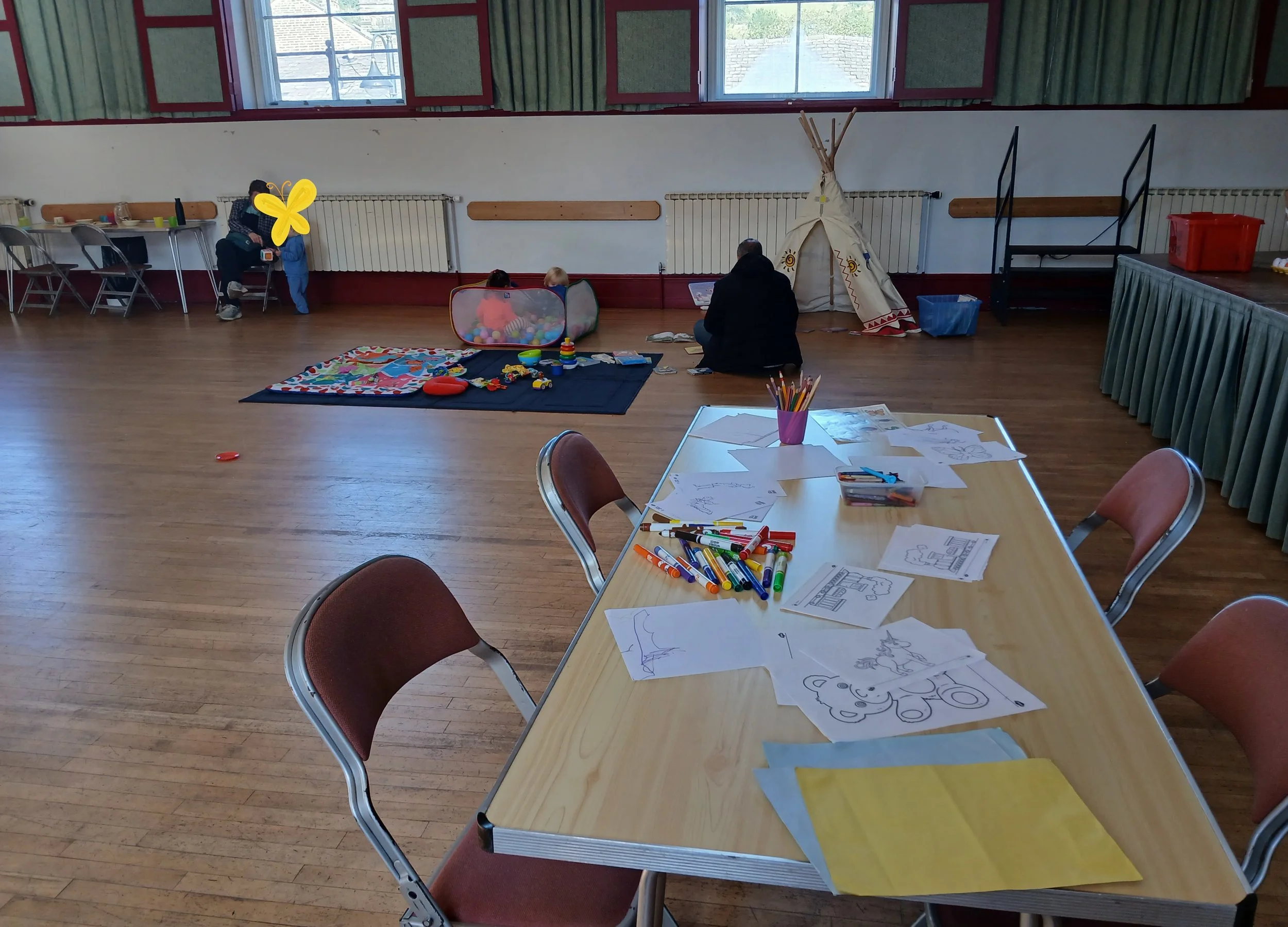 A large room with wooden floors and high windows with green curtains. The room has tables with coloring pages, markers, and coloring supplies. In the background, children and adults are sitting on the floor near a small tent and wall radiators, engaging in activities. One child and an adult are near the windows, one child is playing with a ball, and an adult is reading a book.