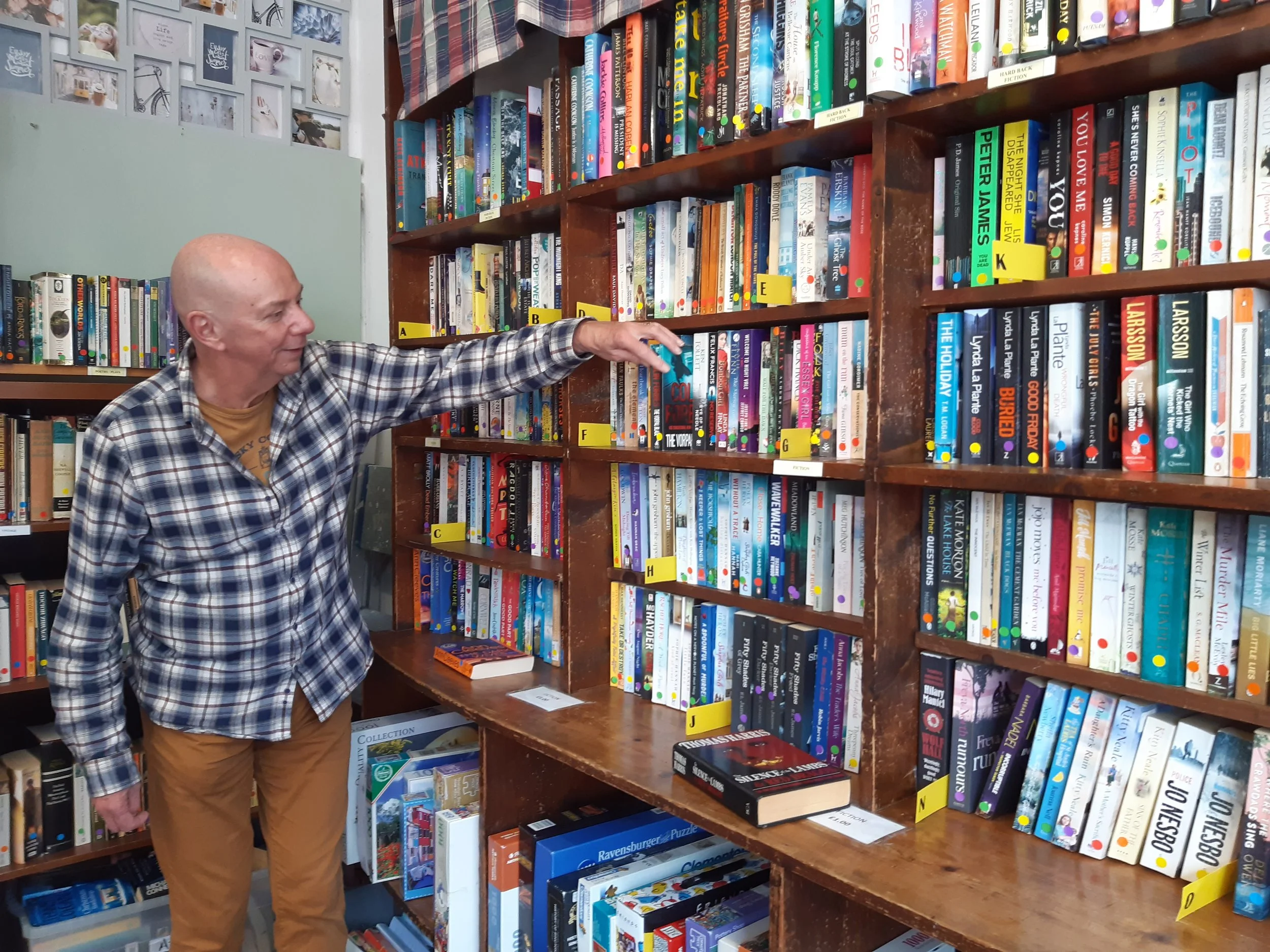 An older man wearing a plaid shirt and beige trousers is browsing books in a bookstore in Maraden, reaching for a book on a wooden bookshelf filled with colourful books.