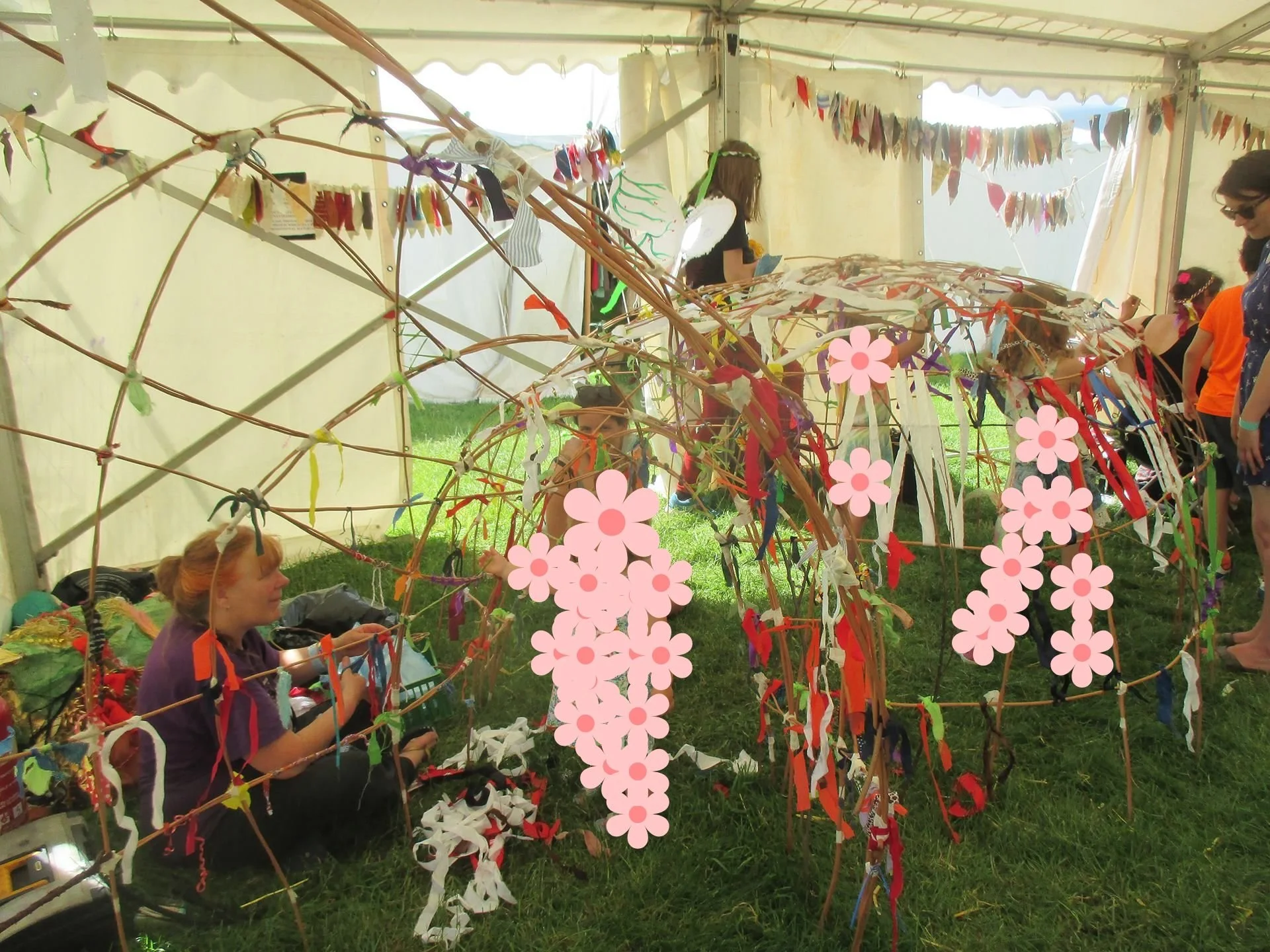 People participating in a colorful outdoor craft activity under a tent, making and decorating a large piece of artwork using ribbons, fabric, and other materials, with bunting hanging above.