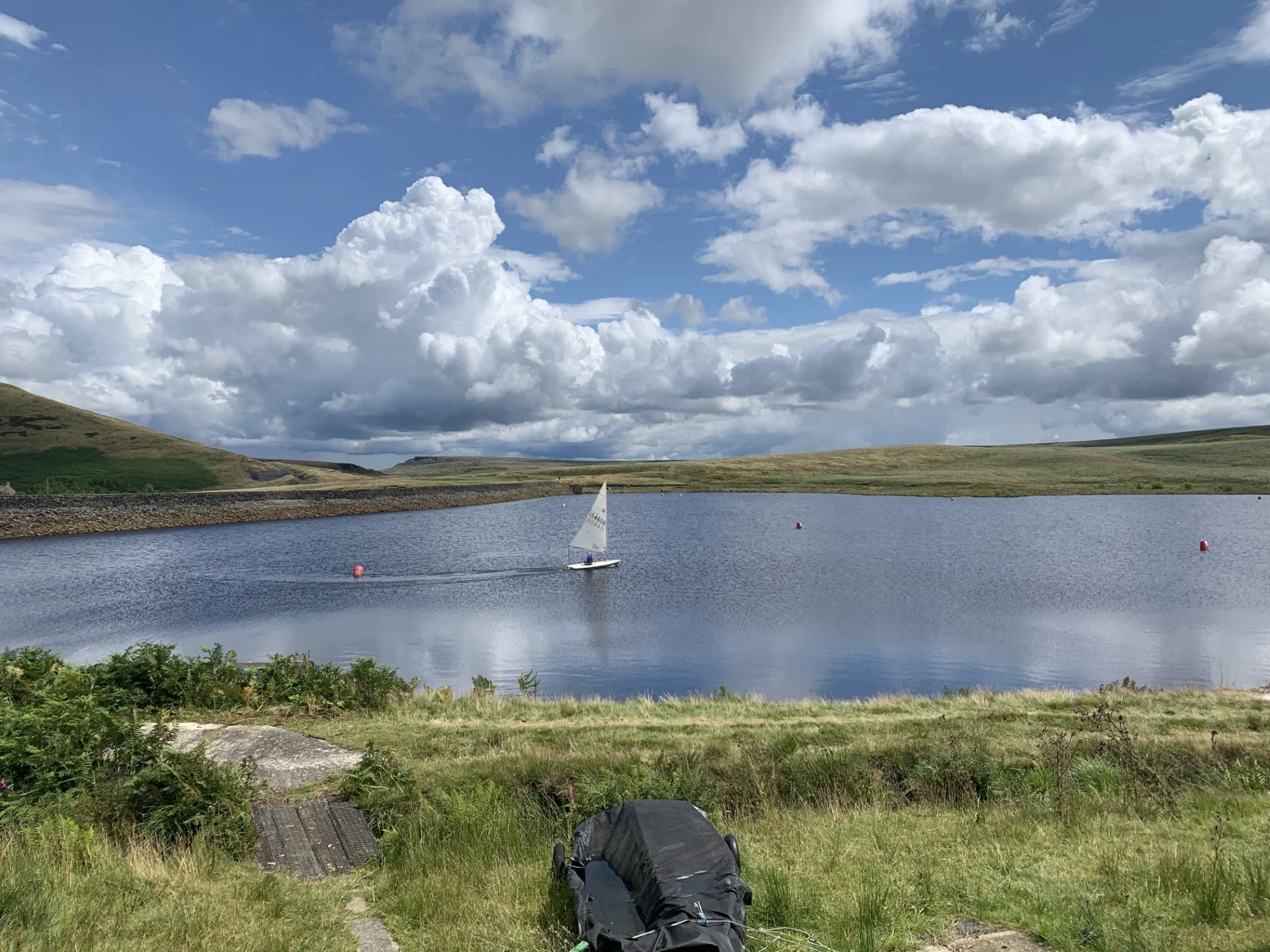 A boat with a sail on a reservoir, surrounded by green hills, under a partly cloudy sky.