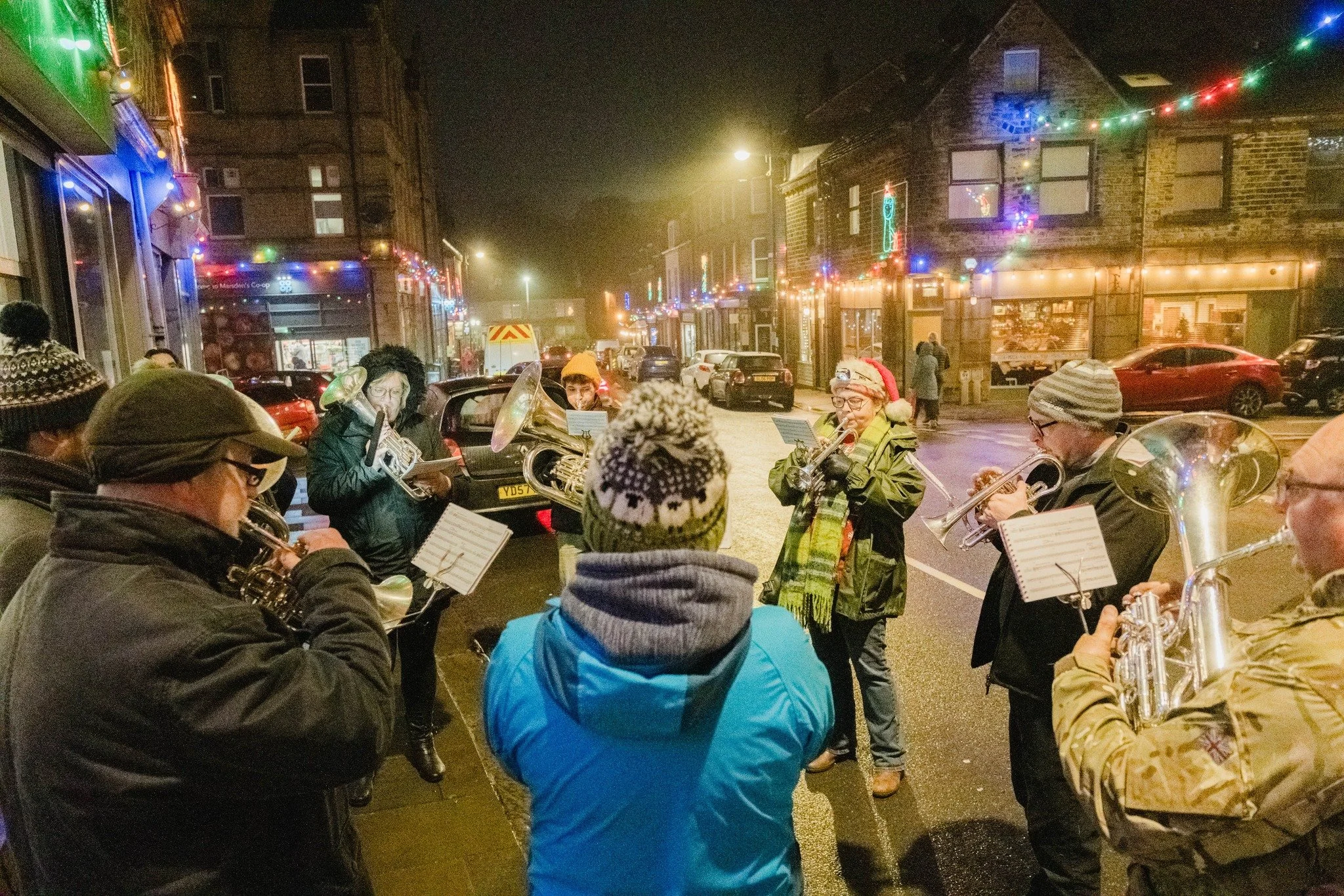 A group of people playing musical instruments, likely part of a band, on Peel Street in Marsden at Christmas. The street is decorated with colourful Christmas lights, with parked cars and buildings in the background.