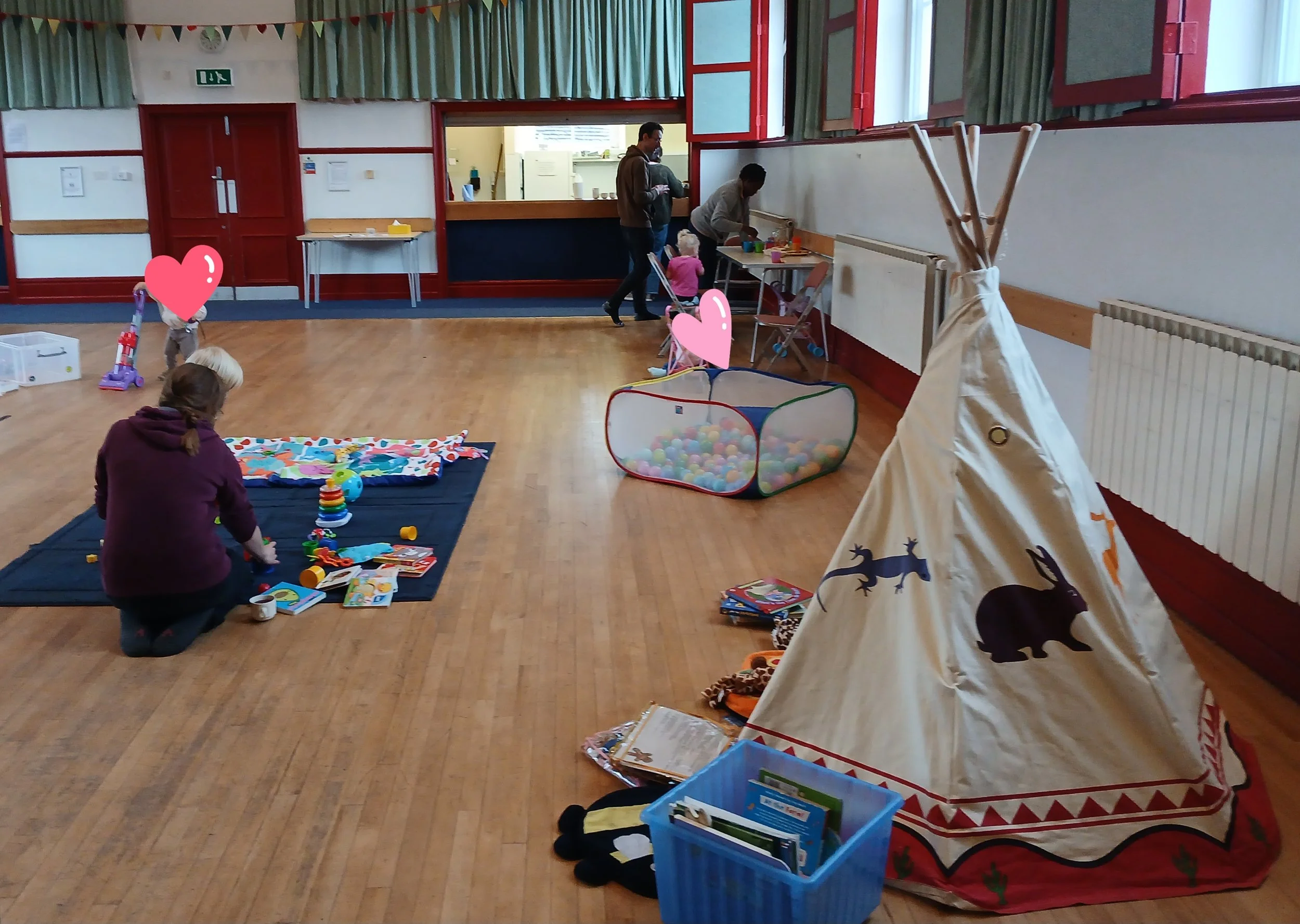 Children and adults in a community center or playroom, engaging in play and activities. There are toys, books, and a ball pit on the floor, with a wooden teepee tent on the right. The room has a wooden floor, red and green window frames, and a service counter with people in the background.