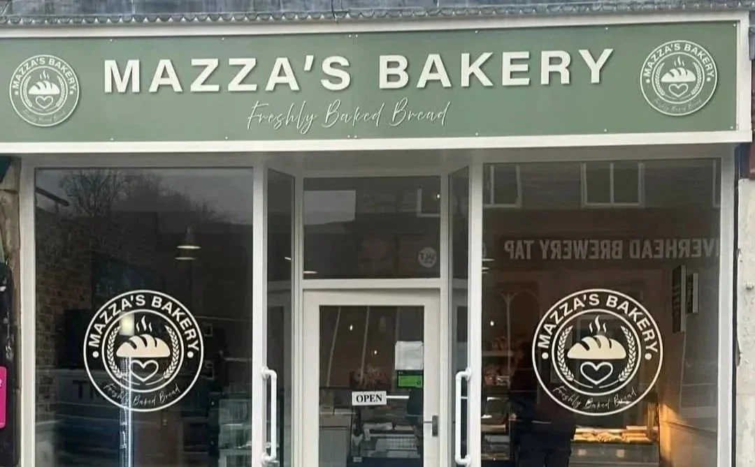 The storefront of Mazzas Bakery with a green sign that reads 'Mazza's Bakery' and 'Freshly Baked Bread.' The glass doors feature the bakery's logo with a steaming loaf of bread and a heart, along with the text 'Mazza's Bakery, Freshly Baked Bread.' Inside, there are visible interior elements and a window with a sign that says 'OPEN.'