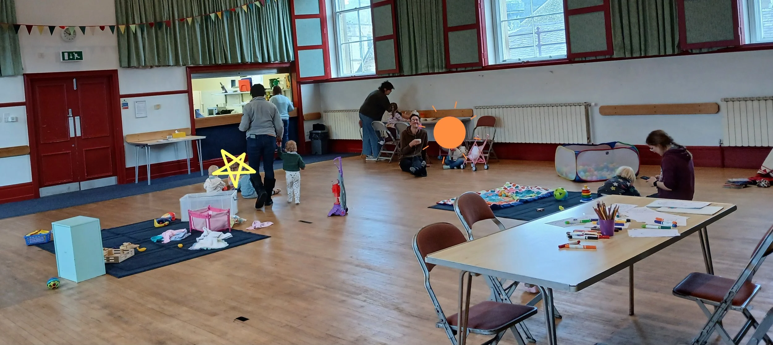 A playroom with kids and adults. Toys, markers, and art supplies on tables and mats. Children playing with toys and interacting with adults. Large windows letting in natural light. Chairs and tables arranged across the room.