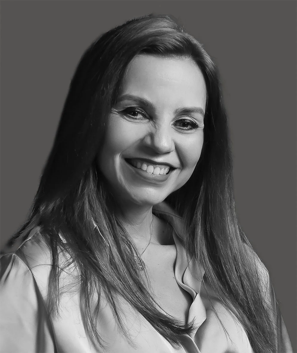 Retrato em preto e branco de uma mulher sorridente com cabelo longo e escuro, usando blusa clara e fundo cinza.