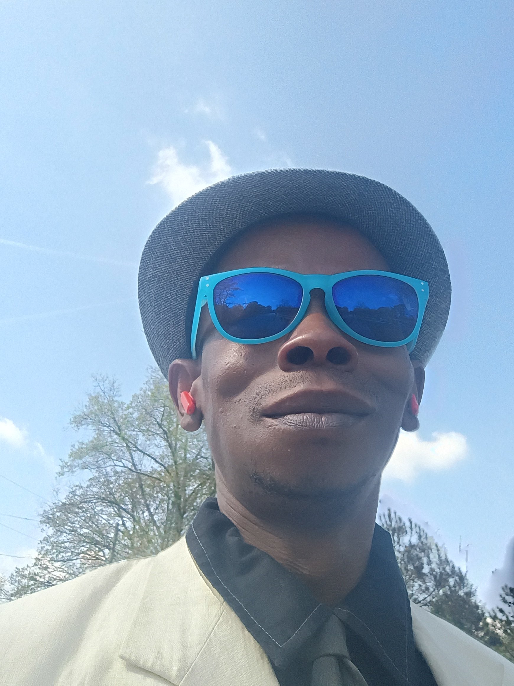 A person wearing a gray hat, blue sunglasses, pink earrings, and a beige blazer is outdoors with blue sky and trees in the background.