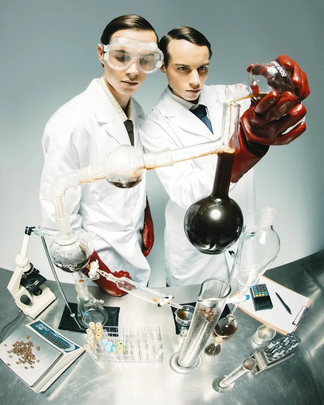 Two scientists wearing white lab coats, goggles, and red gloves conducting a chemistry experiment with glassware containing dark liquids, on a metallic laboratory table.