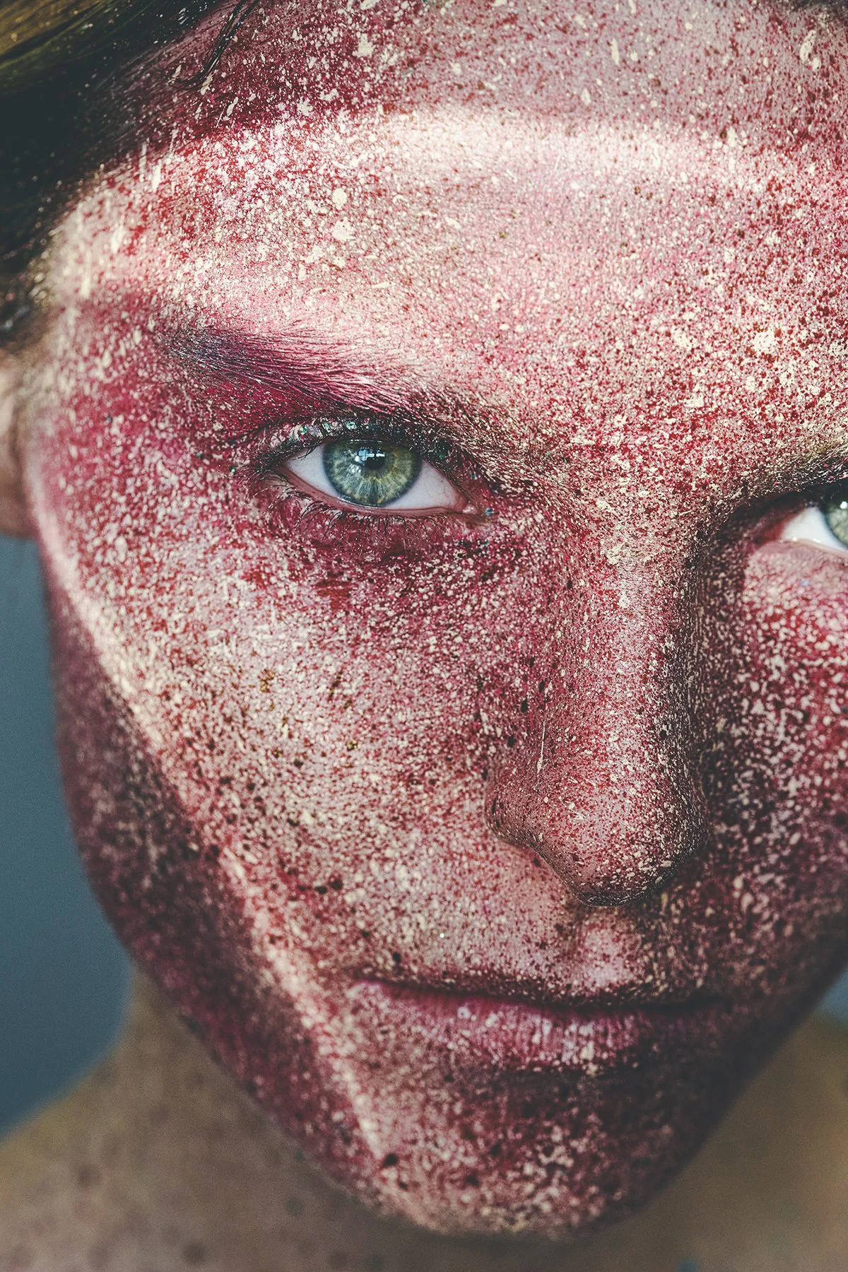 Close-up of a woman's face covered in red and gold glitter, with striking blue eyes and partially closed lips.