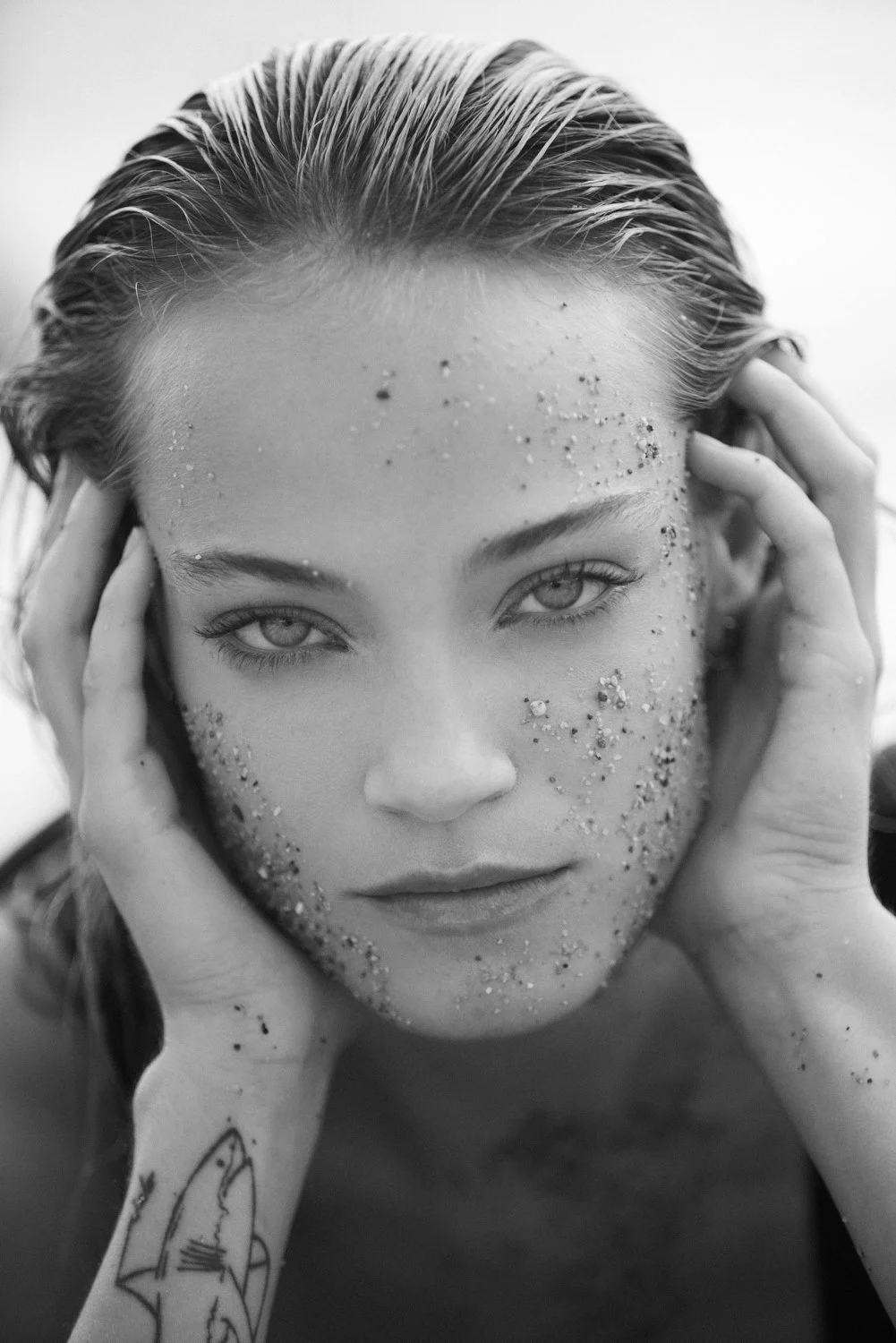 Black and white close-up of a woman with wet hair, hands on her head, and glitter on her face, looking directly at the camera.