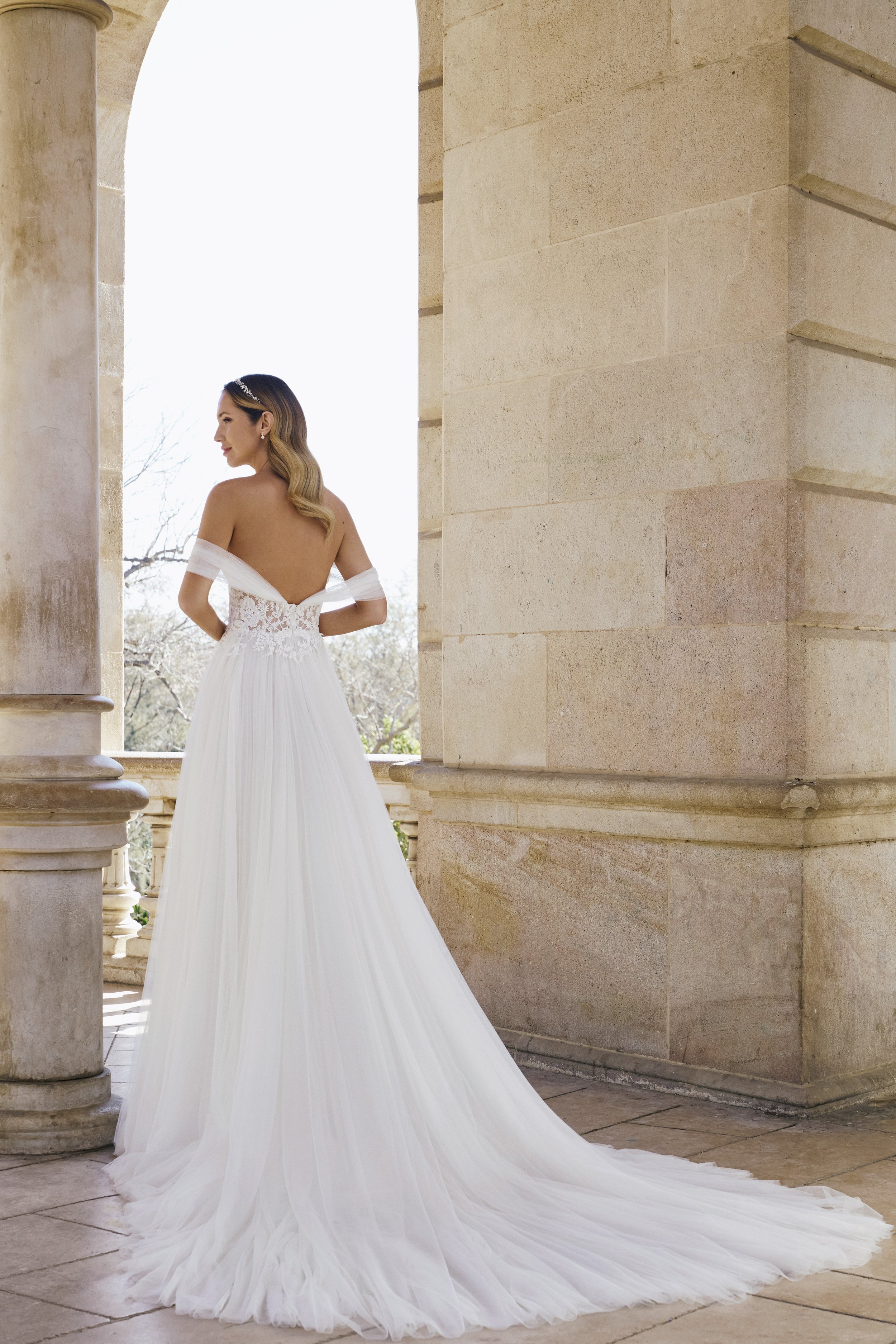 A bride in a white wedding gown with off-the-shoulder sleeves, standing on a stone terrace with columns, looking to the side with trees visible in the background.