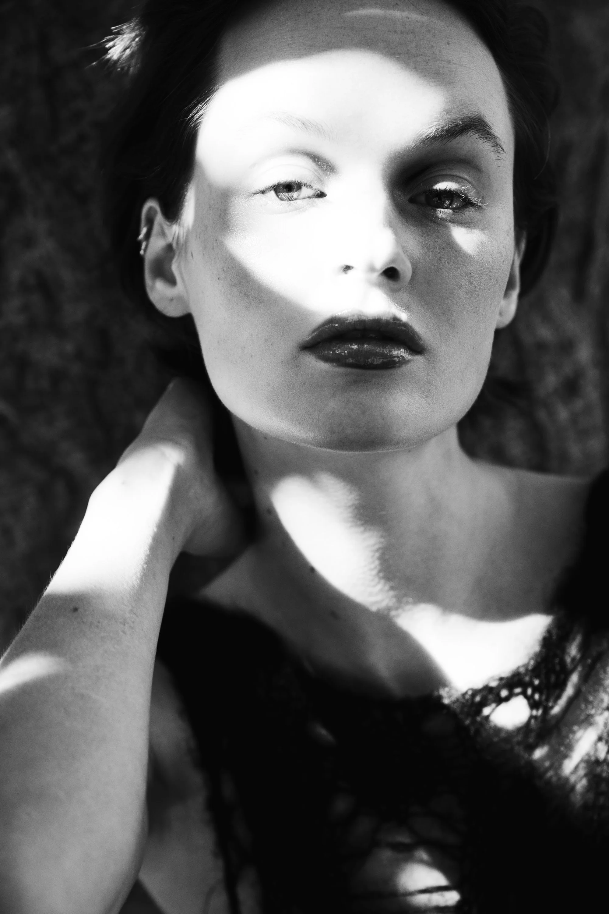 Black and white photo of a woman lying down, with sunlight casting shadows across her face, wearing dark lipstick and lace clothing.