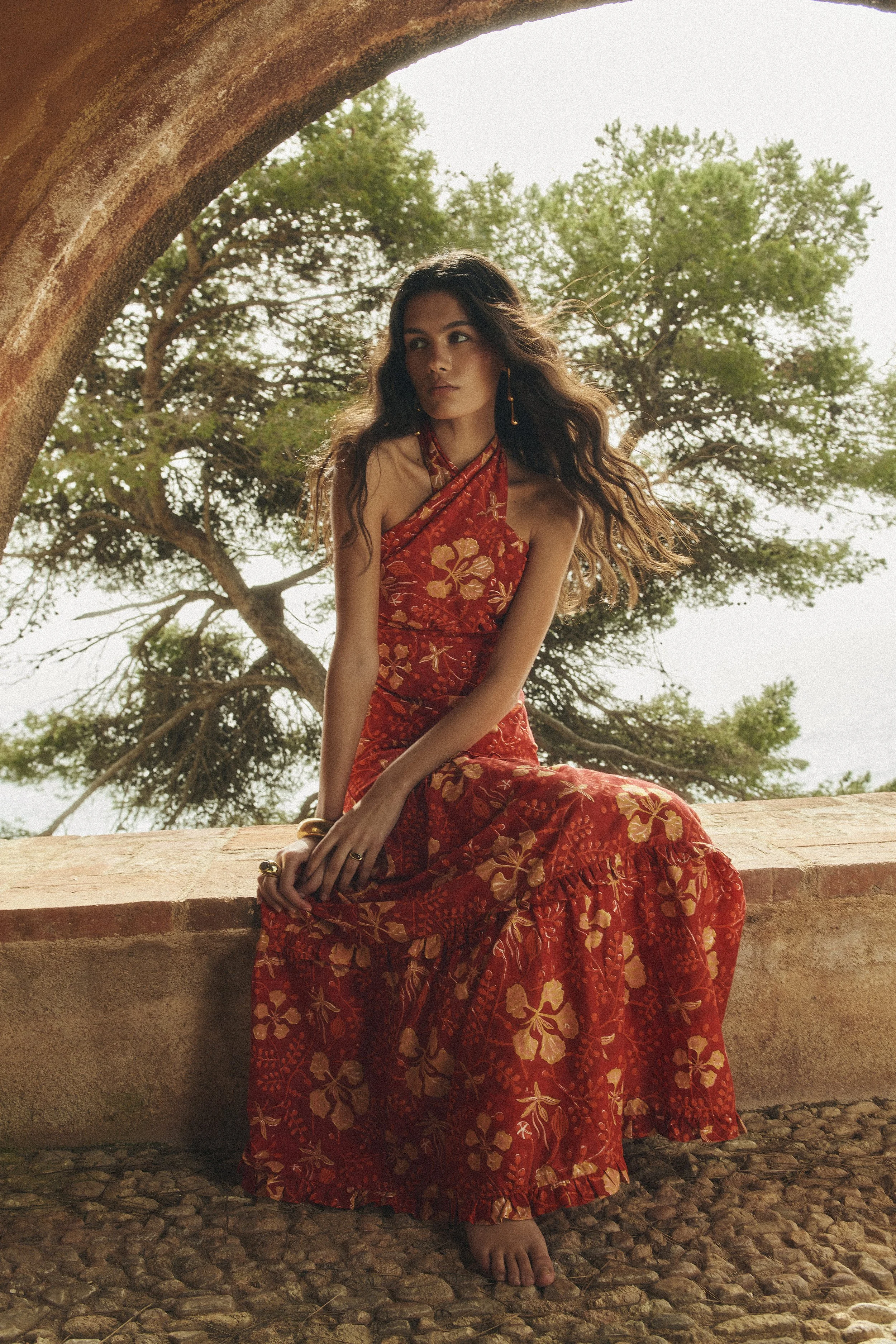 A woman in a red floral dress sitting outdoors on a stone ledge, with trees in the background.