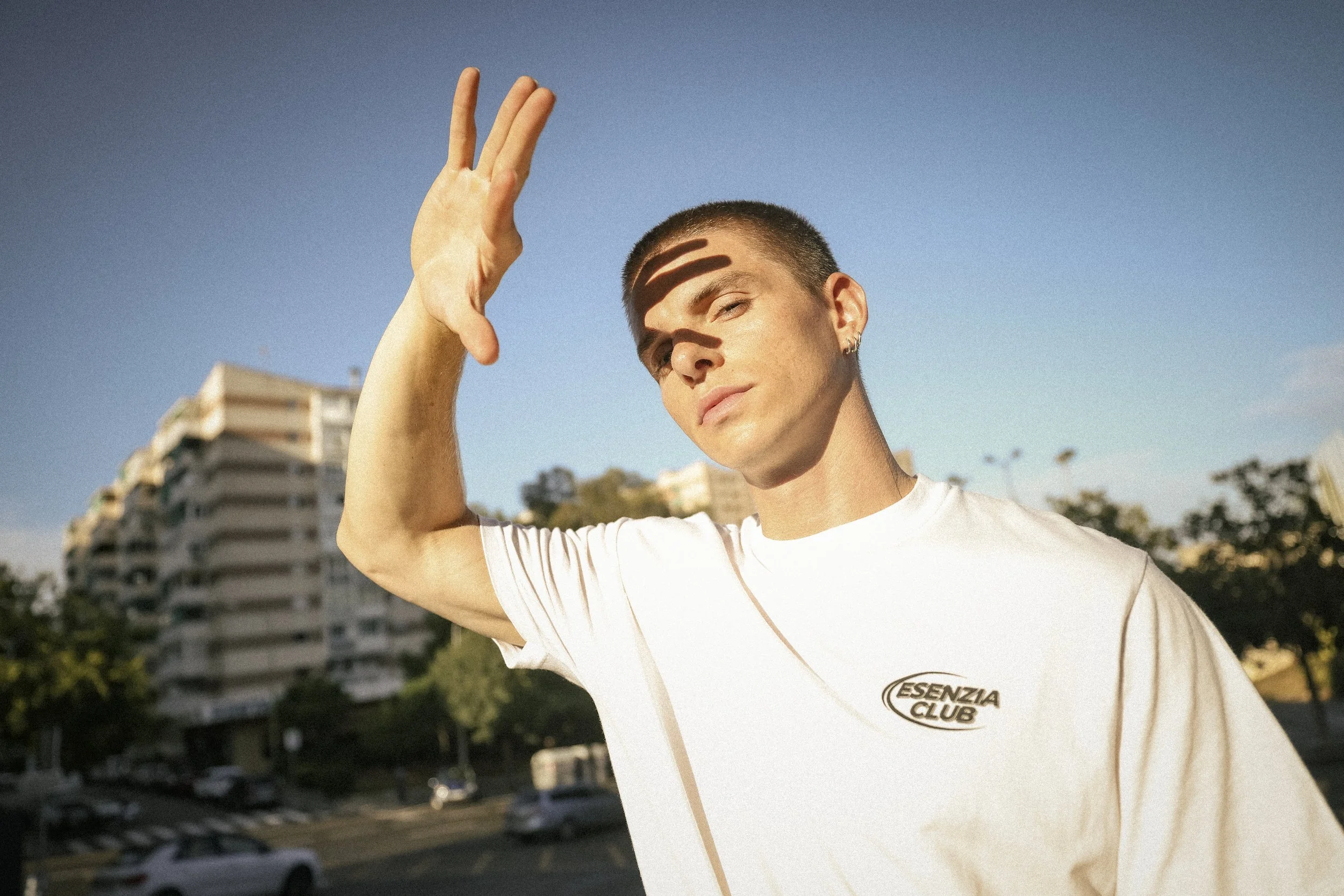 Young man with short hair and earrings raising hand in sunny outdoor urban area with tall building and trees in background