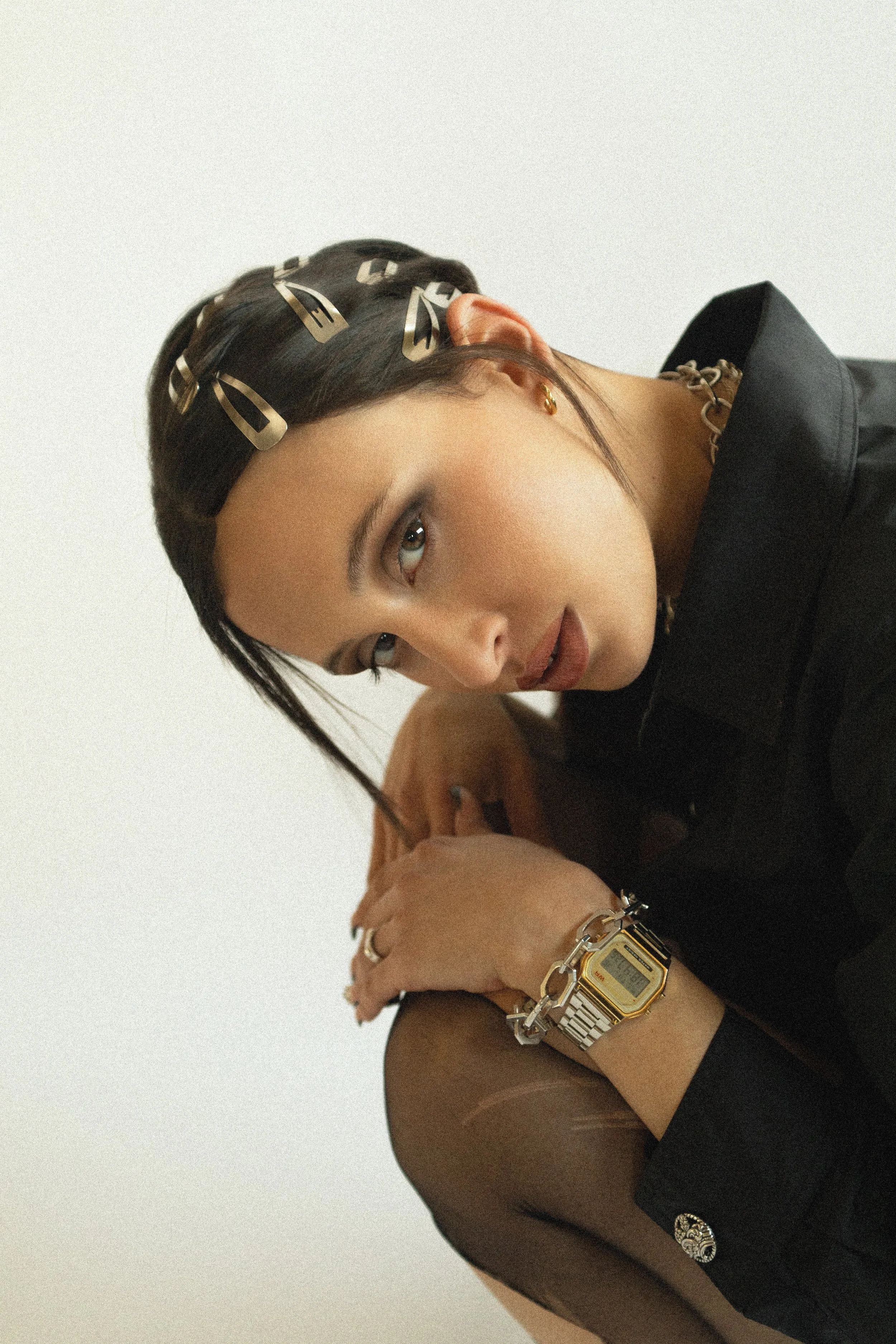 A woman with dark hair styled with gold hair clips, wearing gold jewelry and a black jacket, posed against a plain white background.