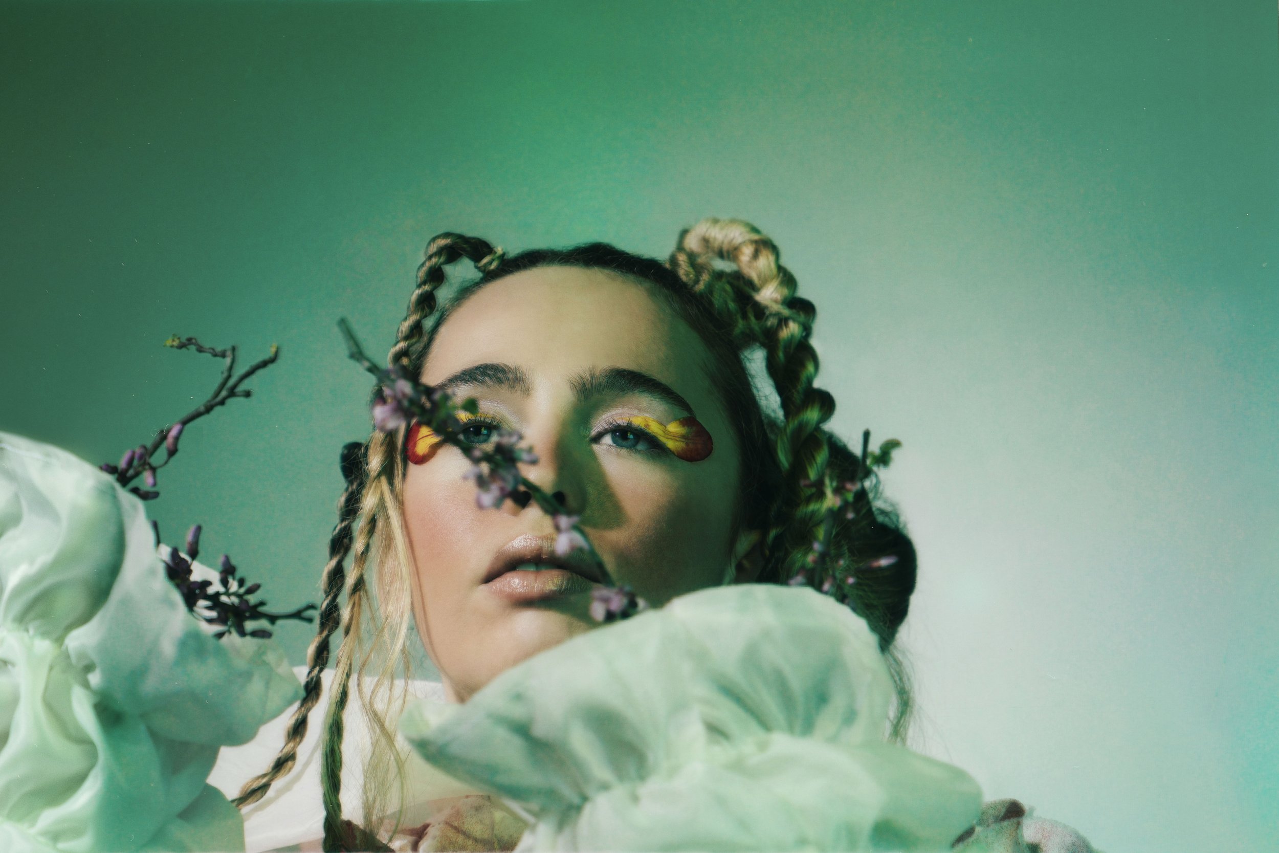 A young woman with braided hair and colorful face makeup, partially obscured by cherry blossom branches, against a green background.