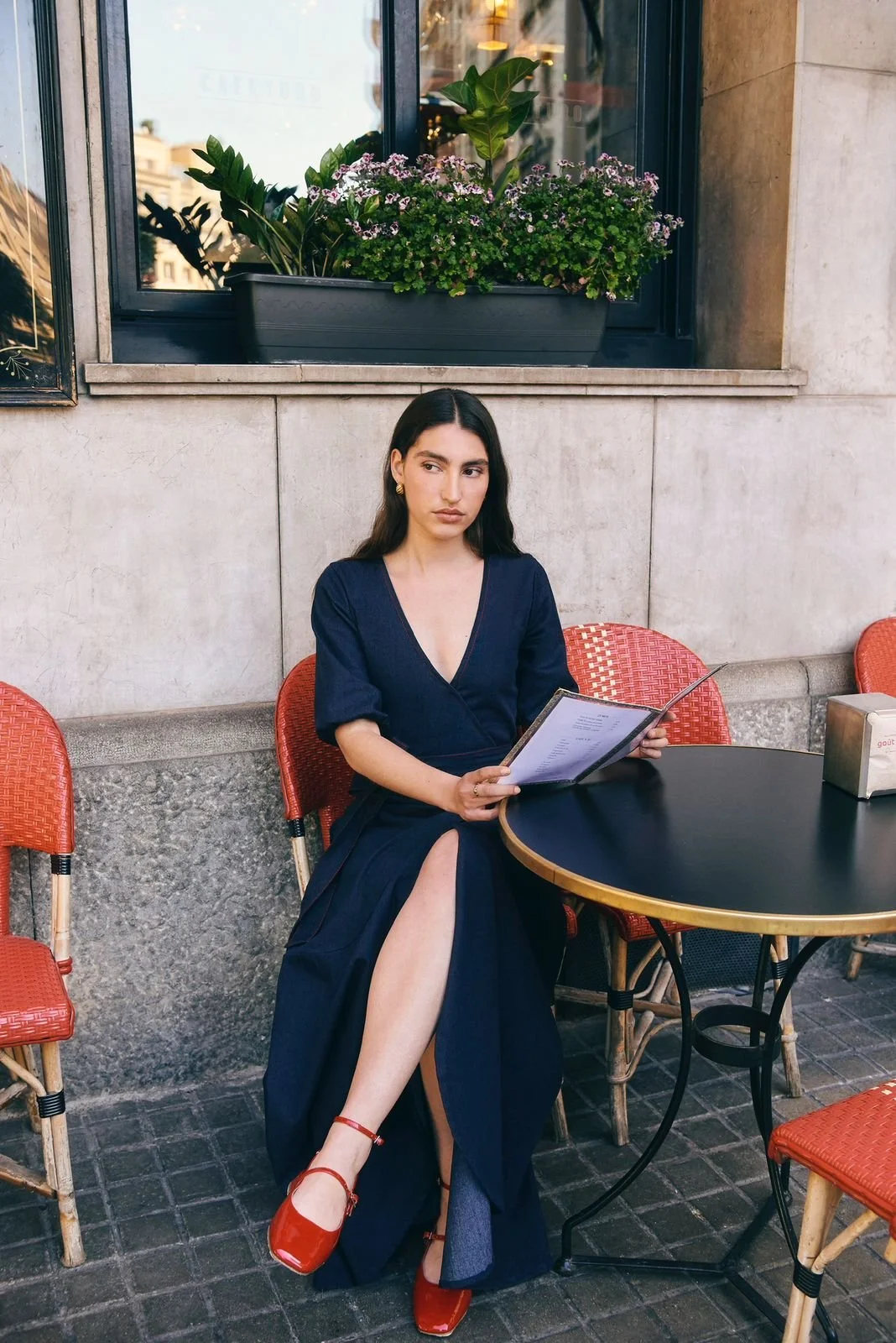A woman sitting at an outdoor cafe table, wearing a navy blue dress with a high slit, red shoes, and accessorized with earrings, holding a menu. There are red chairs around her and a large planter with flowers and green plants on the wall behind her.