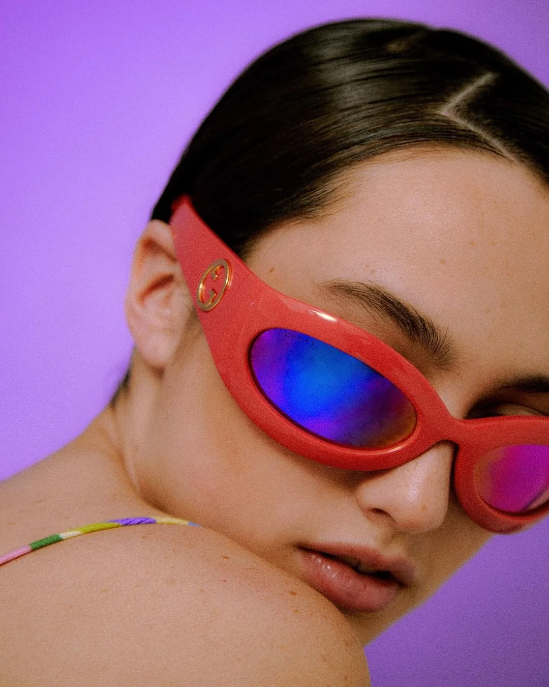 Close-up of a woman with dark hair in a sleek hairstyle wearing red sunglasses with blue mirrored lenses, against a purple background.