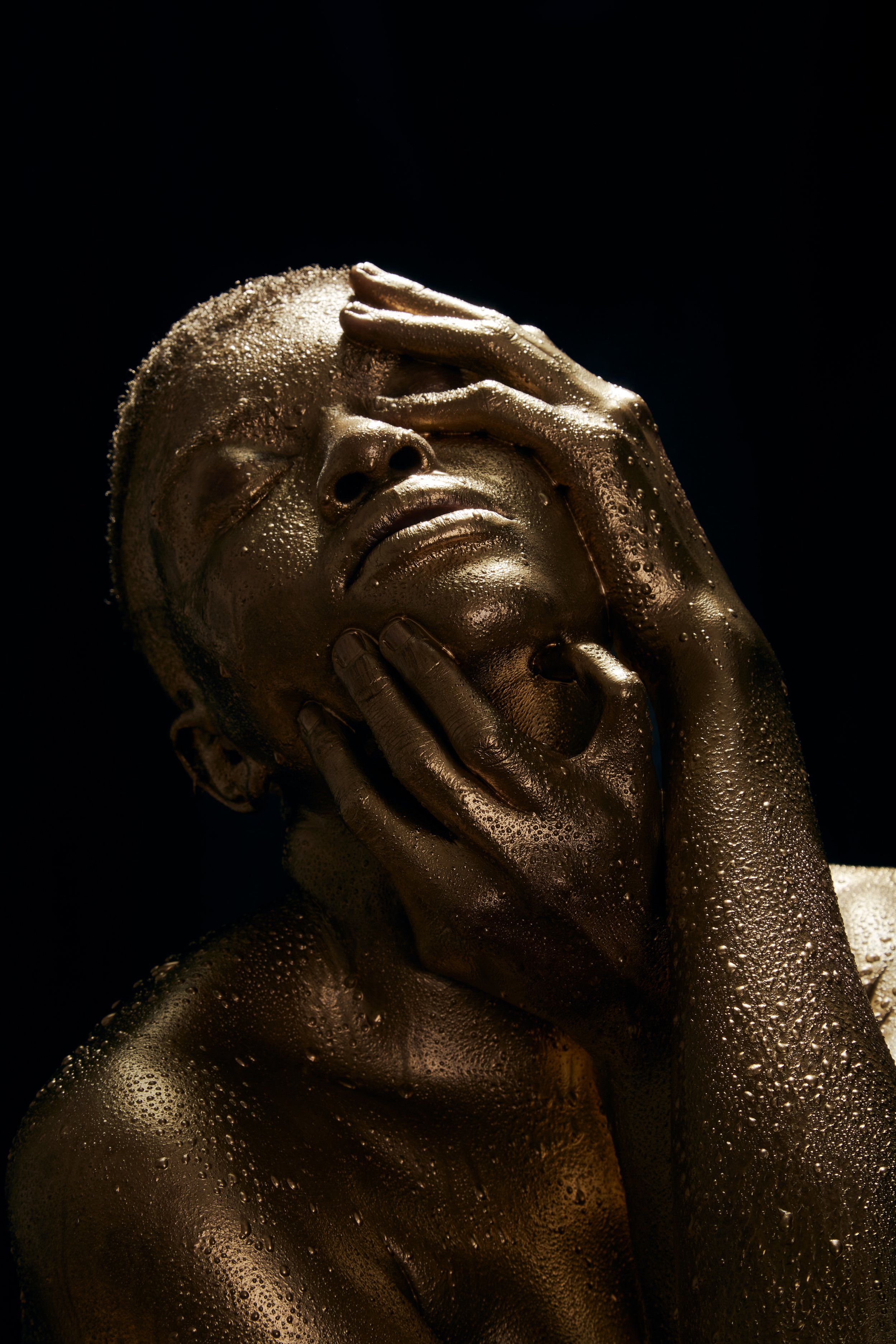 A person with gold body paint and bubbles on their skin, holding their face with one hand and their chin with the other, against a dark background.