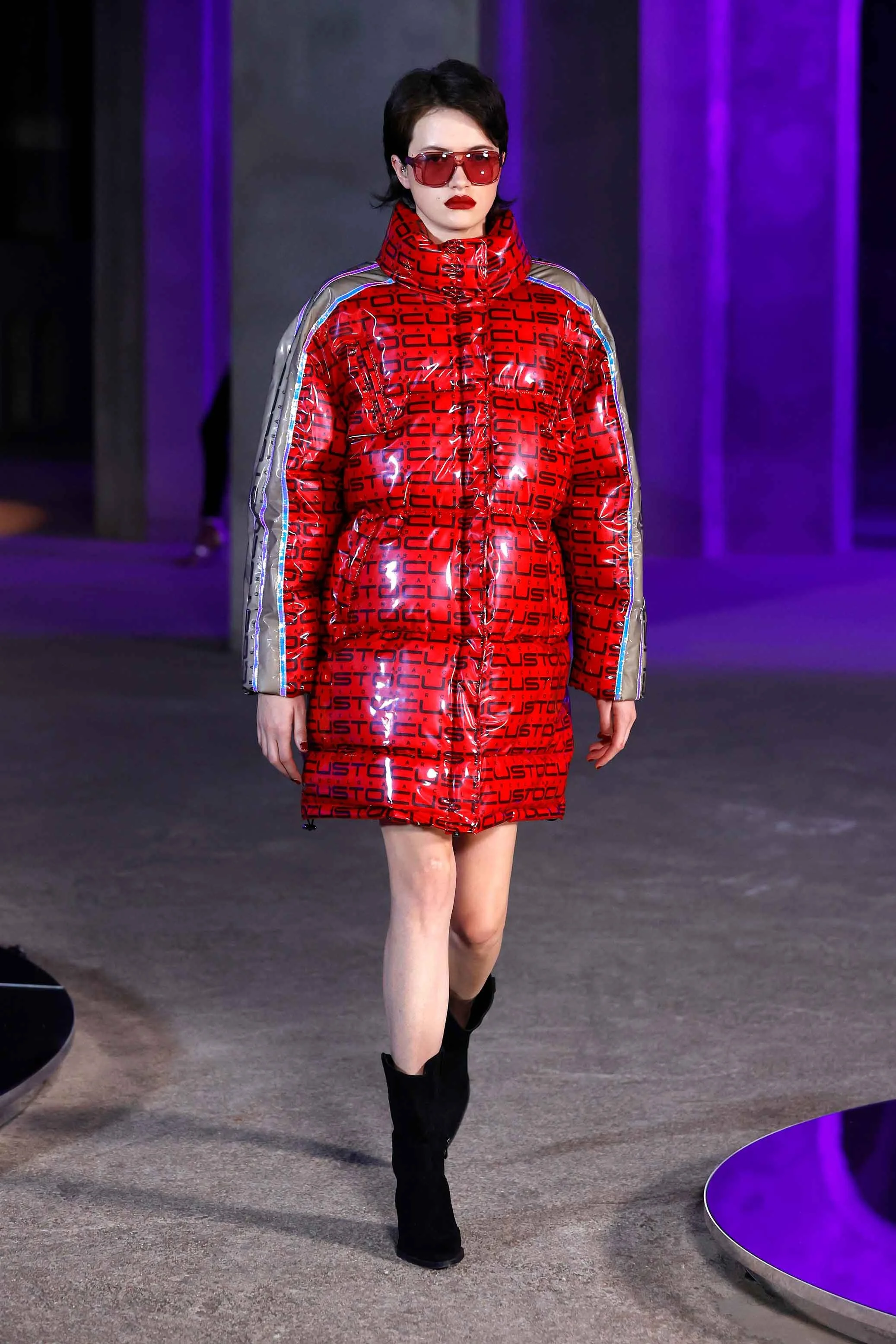 A female model walking on a runway wearing a shiny red jacket with black lettering and silver accents, dark sunglasses, and black boots, with purple lighting in the background.
