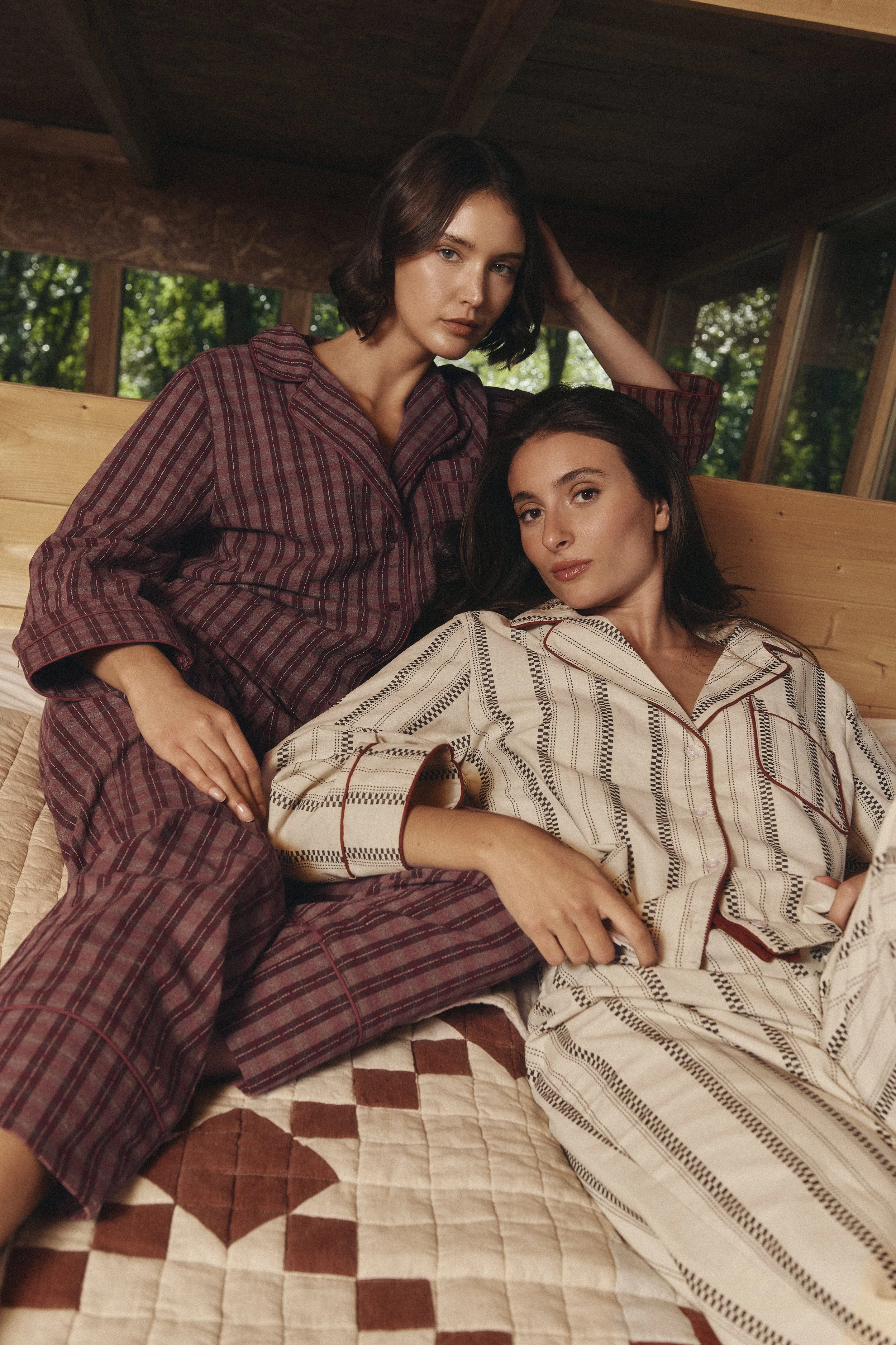 Two women in pajamas relaxing on a bed in a wooden cabin with large windows showing trees outside.