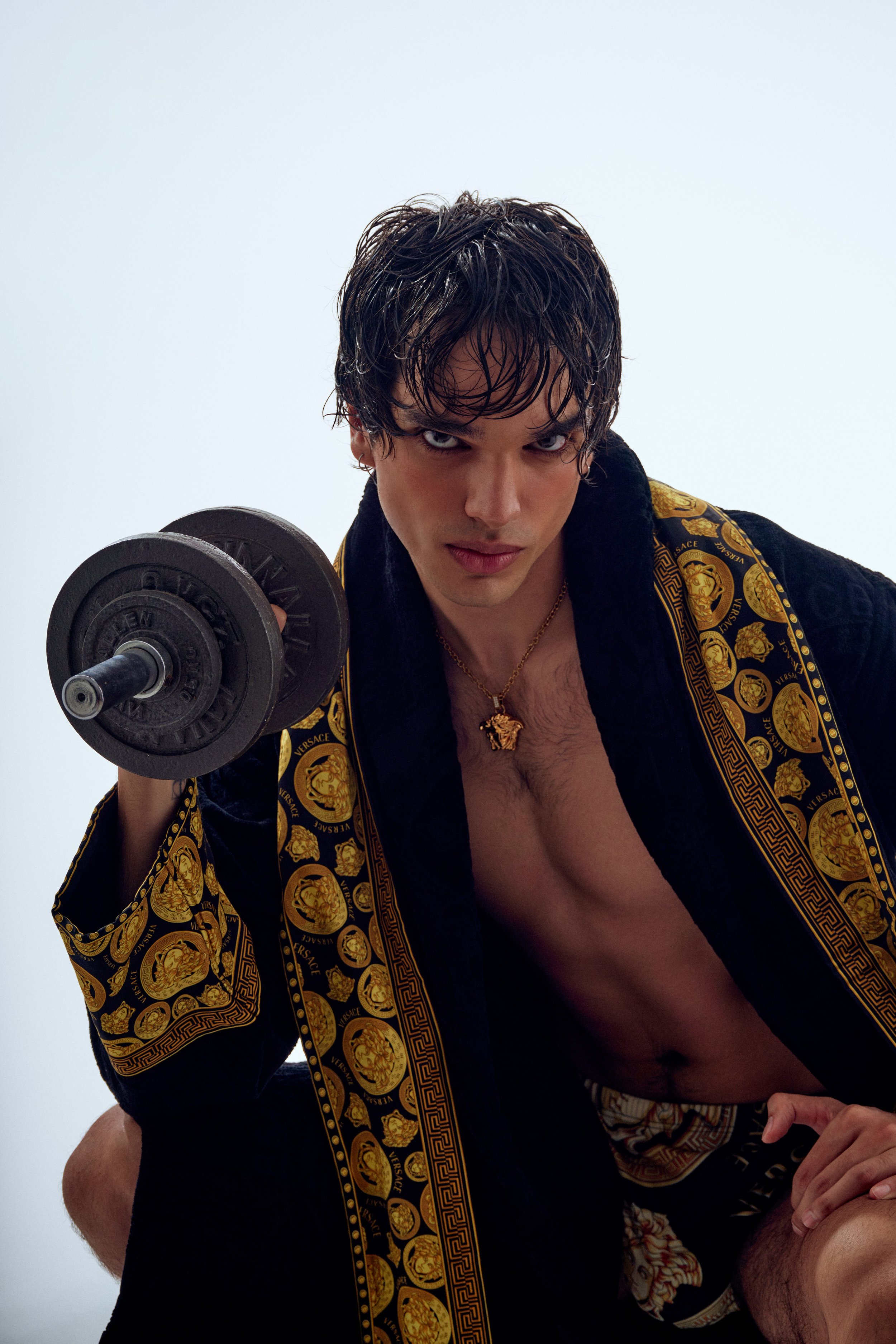 A young man with dark curly hair, wearing a black and gold Versace robe and gold necklace, holding a dumbbell while looking into the camera.