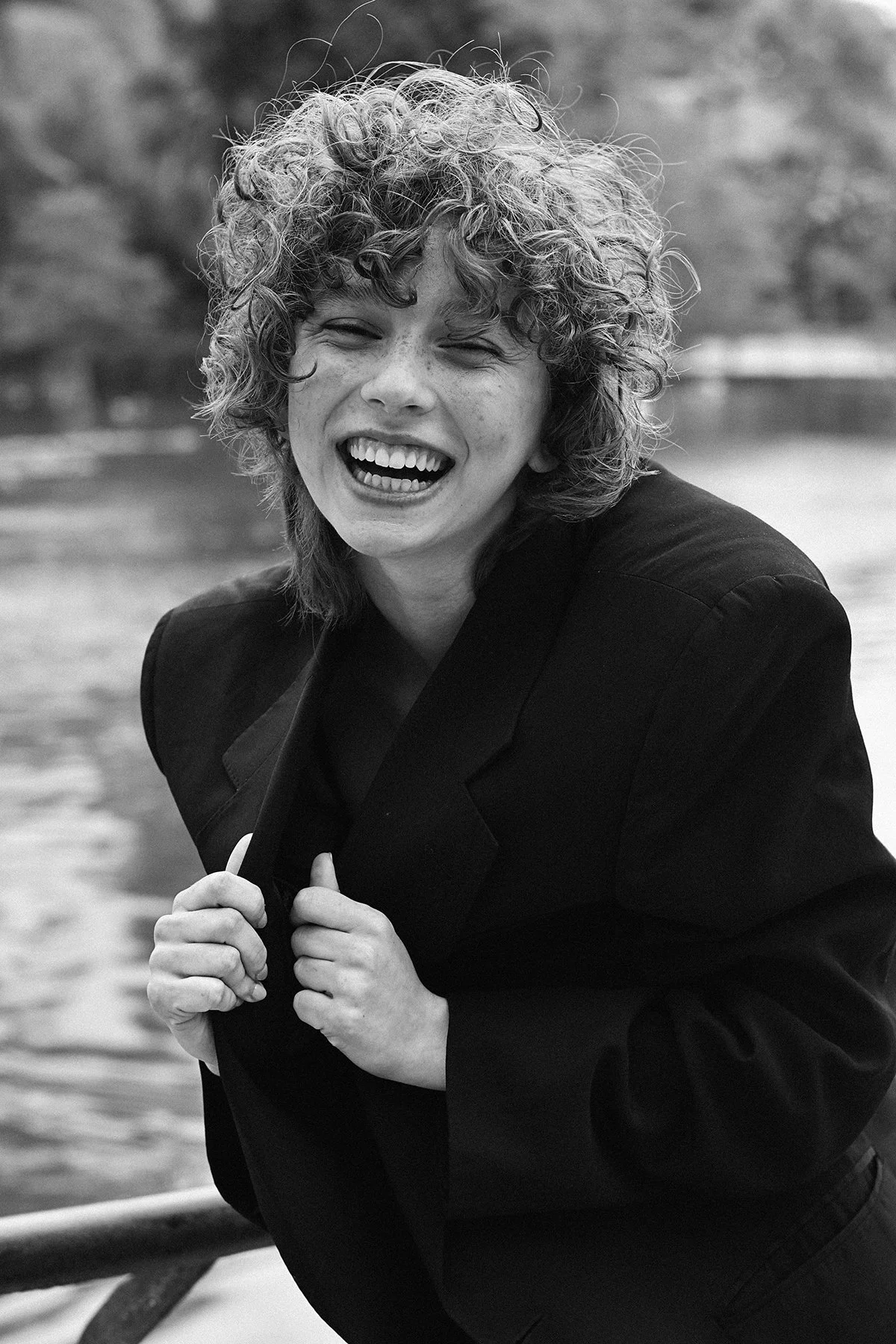 A woman with curly hair laughing and holding her blazer lapels outdoors near a body of water.