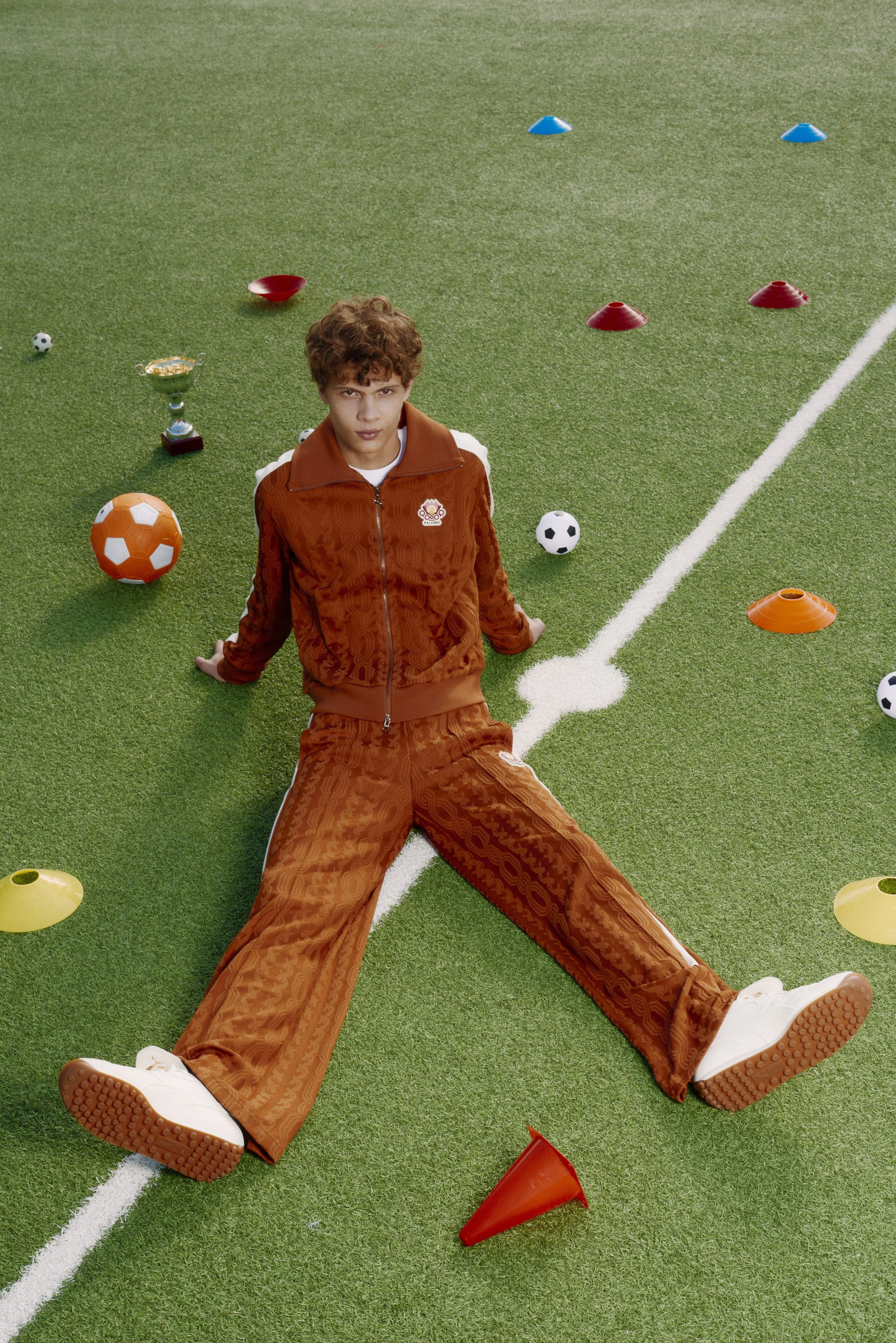 A young man sits on a green turf field, surrounded by soccer balls, small traffic cones, and a trophy, with colored cones scattered around.