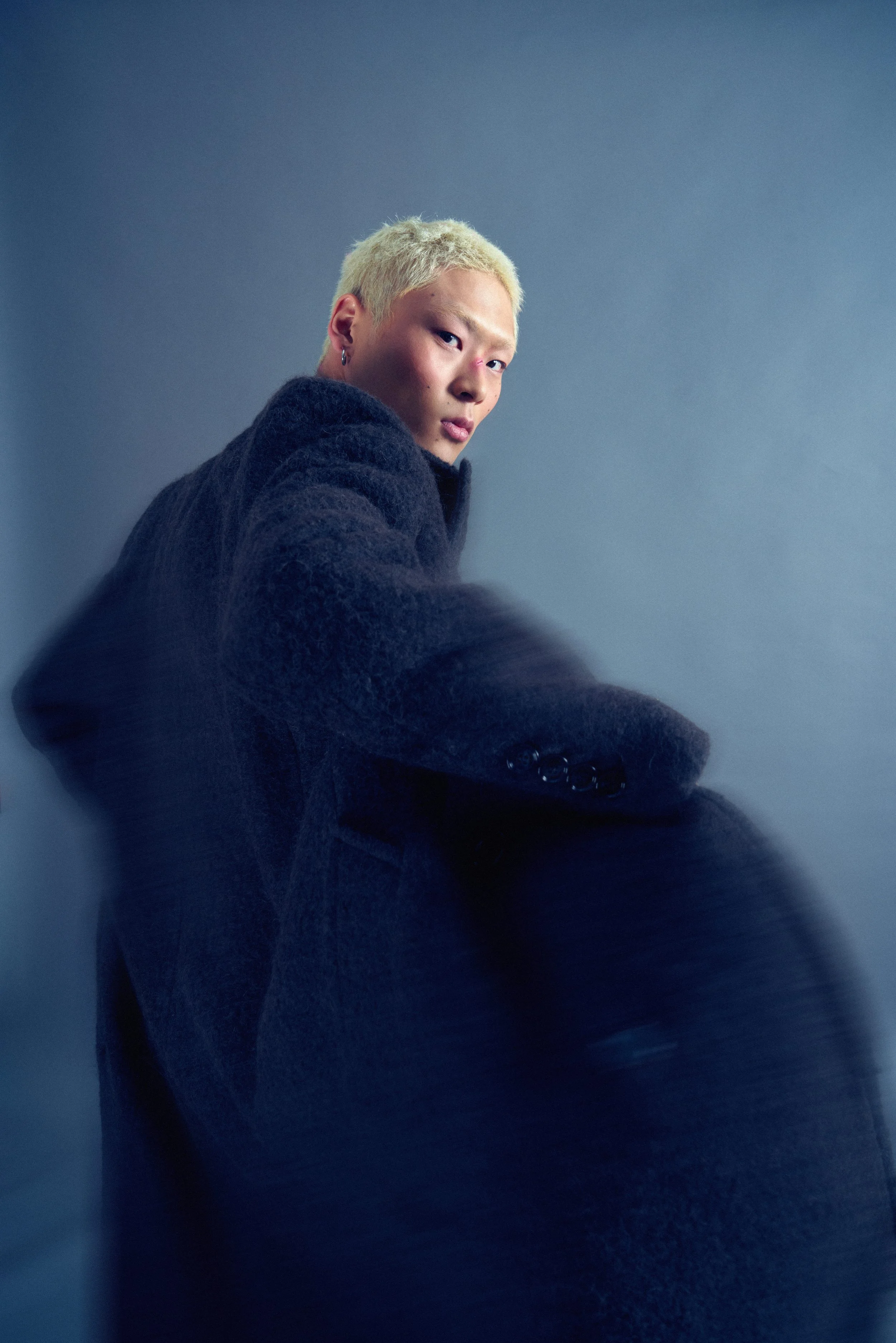 A person with bleached blonde hair, wearing a black textured jacket, looking over their shoulder against a plain gray background.