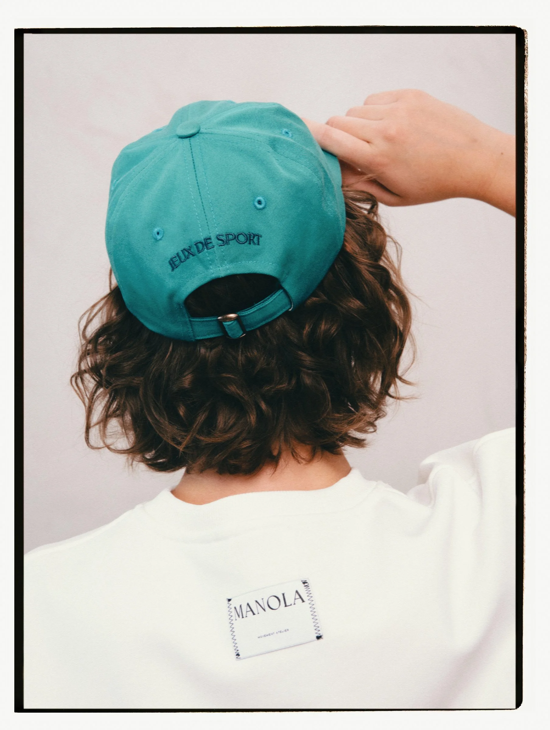 Back view of a person wearing a white Manola t-shirt and a turquoise cap with 'JEUX DE SPORT' embroidered on the back, and their hand touching the cap.