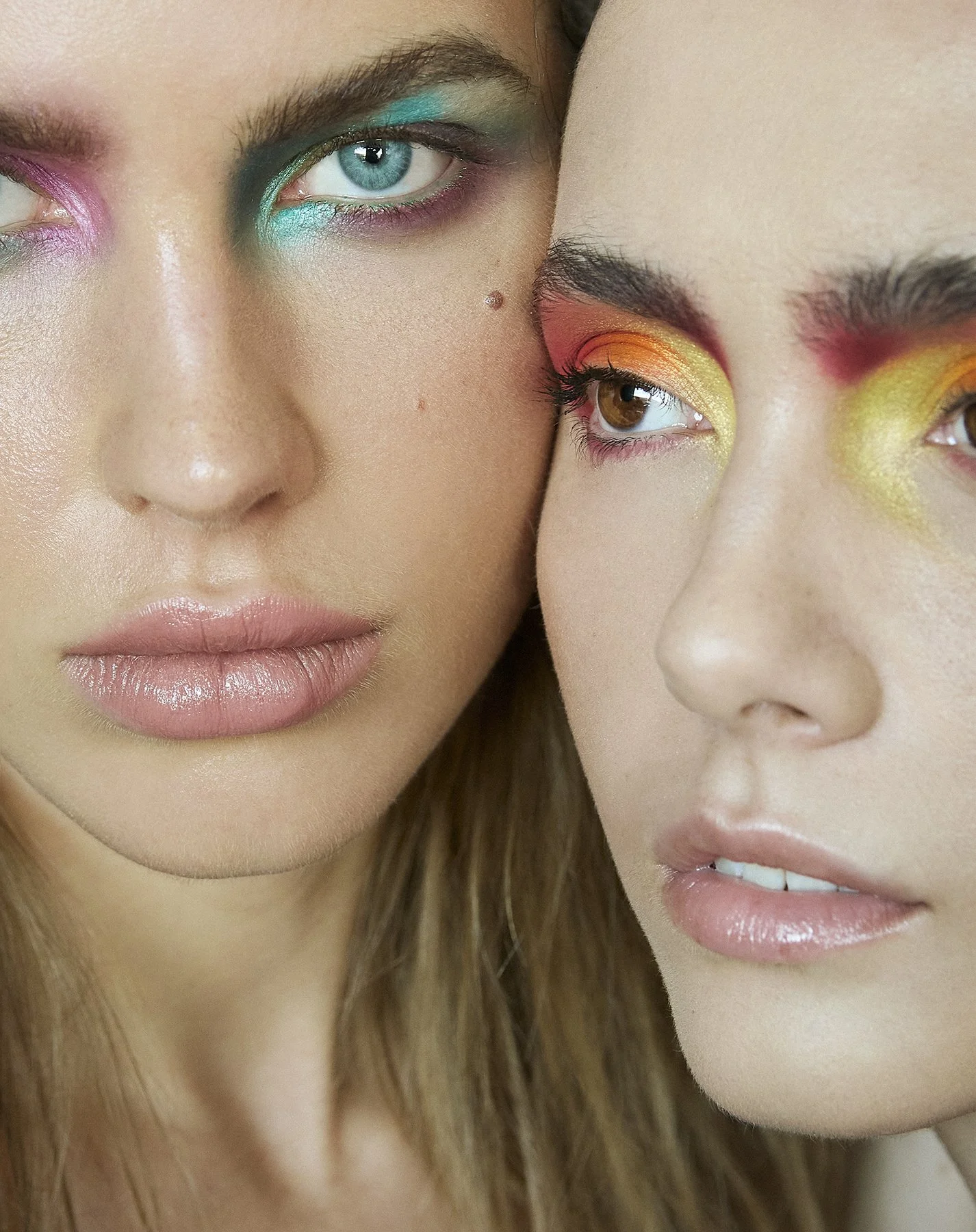 Close-up of two women with colorful, vibrant eye makeup and natural lips, positioned close together.