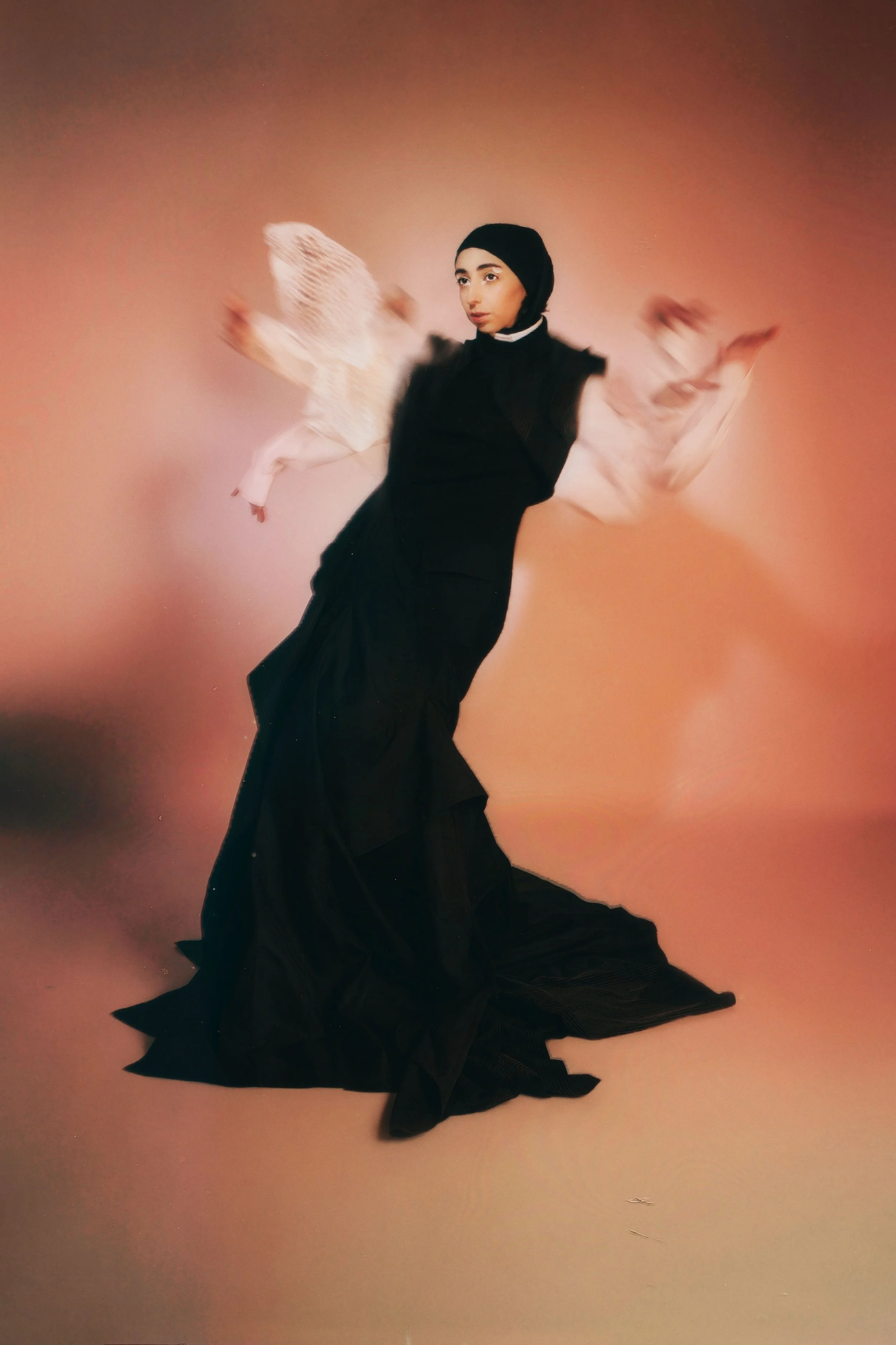 A woman with a serious expression wearing a black hijab and layered black dress, standing against a pinkish-orange gradient background.