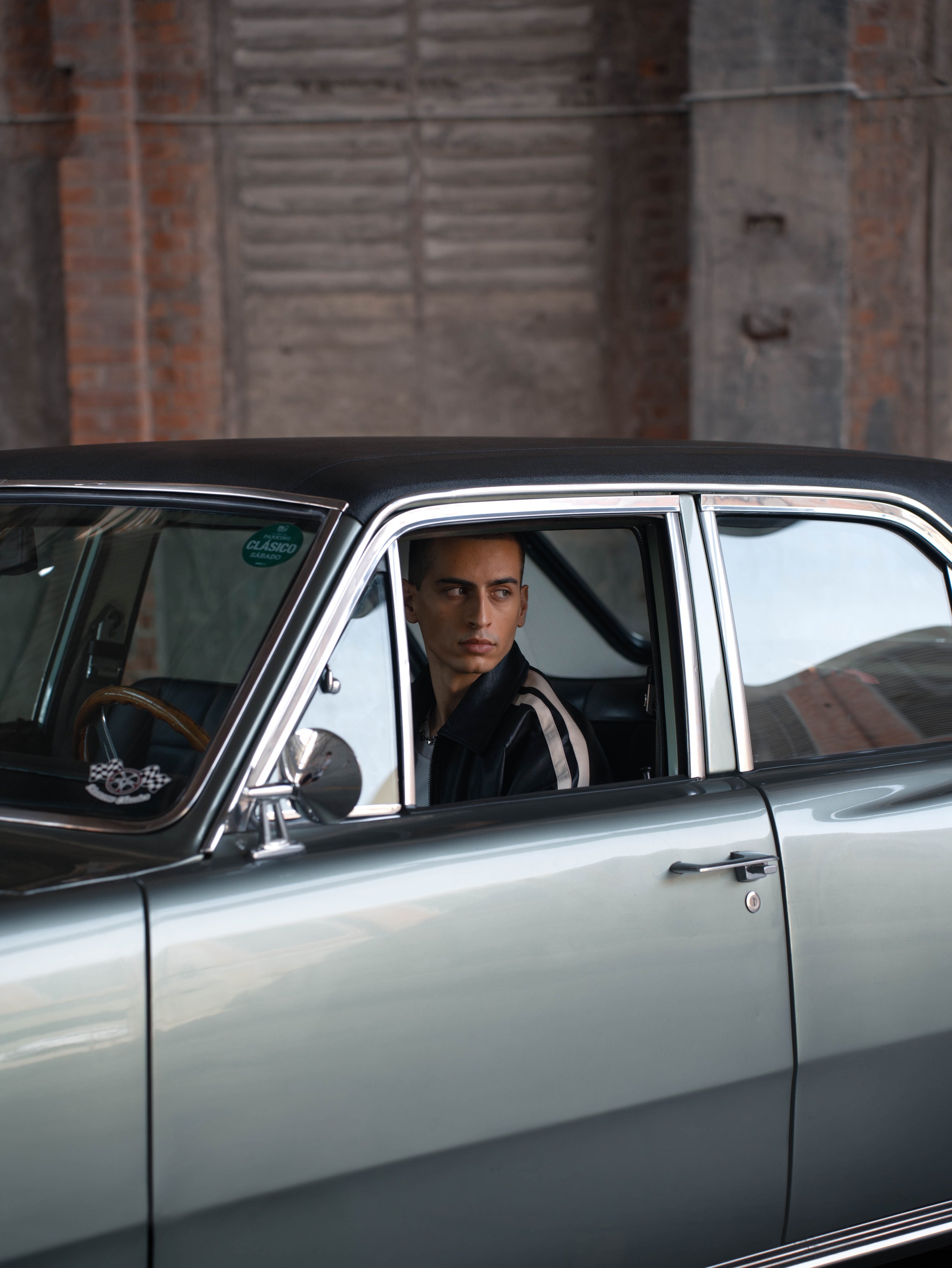 A young man sitting in the driver's seat of a vintage silver car, looking out the window with a serious expression, inside a rustic building with brick and wood walls.