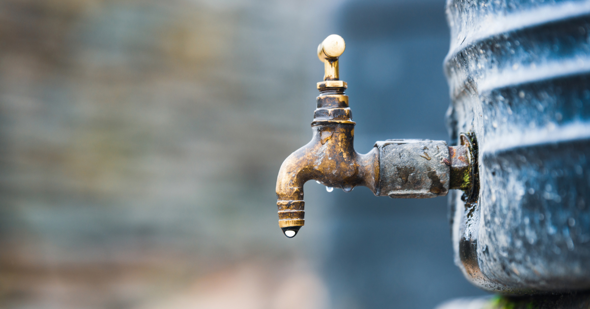 Rusty outdoor water faucet with a droplet of water hanging from the spout, attached to a stained water tank or container.