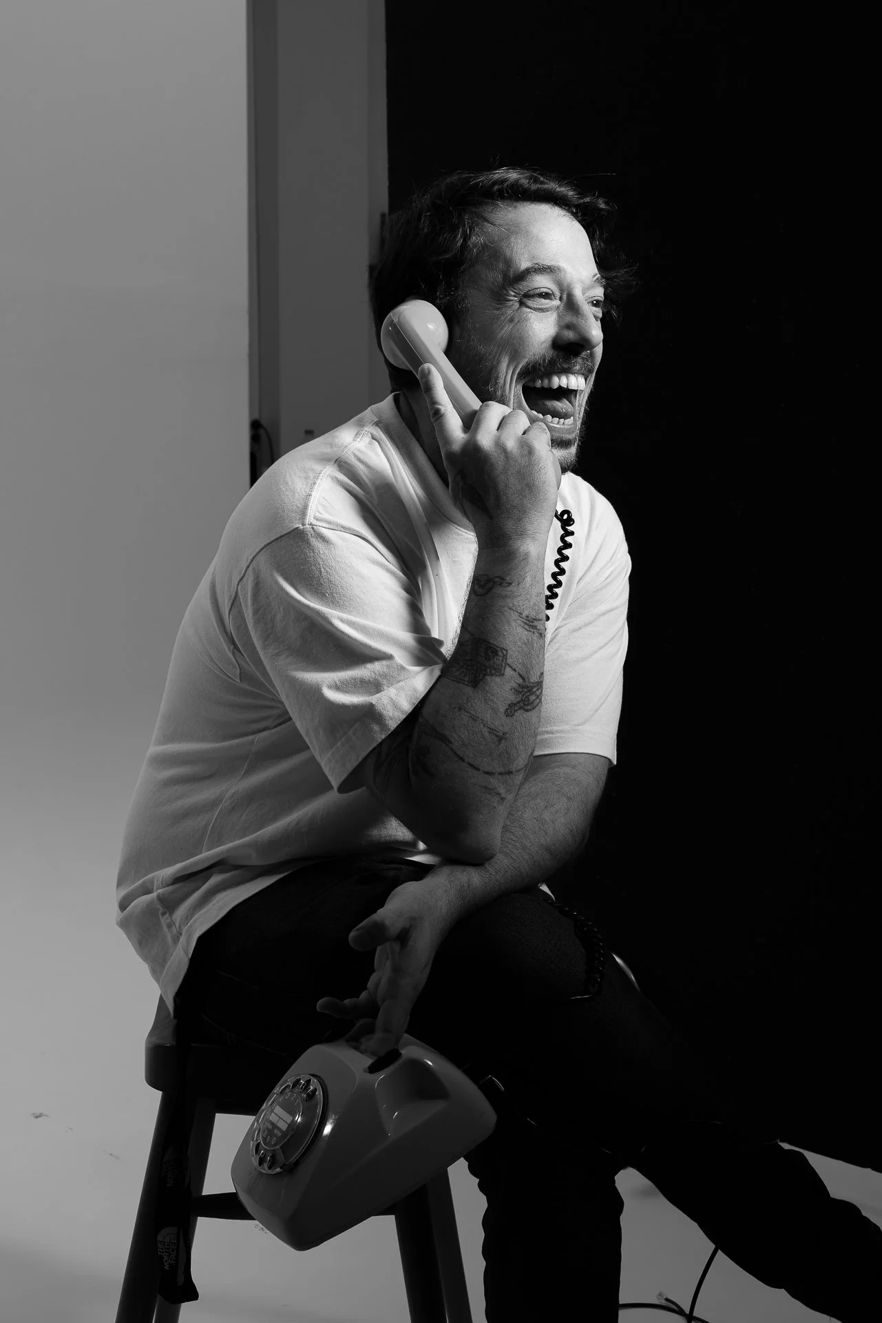Black and white photo of Konstantinos Ntekoumes with tattoos on his right arm, sitting on a stool, talking on a rotary dial phone, laughing, with a dark background for contact infos