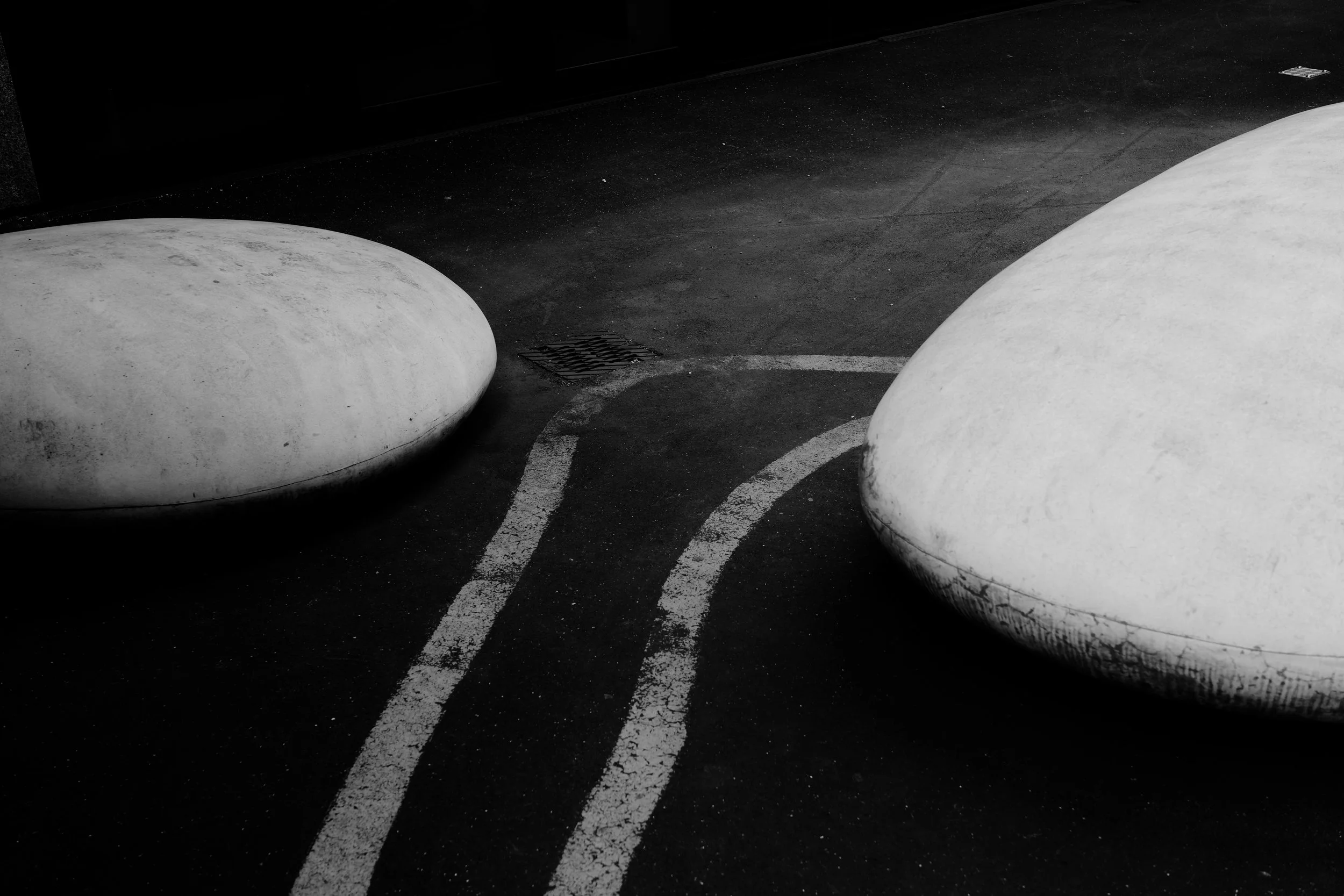 Zwei große, runde, weiße Betonbänke auf einer asphaltierten Oberfläche mit weißen Linien, im Hintergrund dunkle Wand.