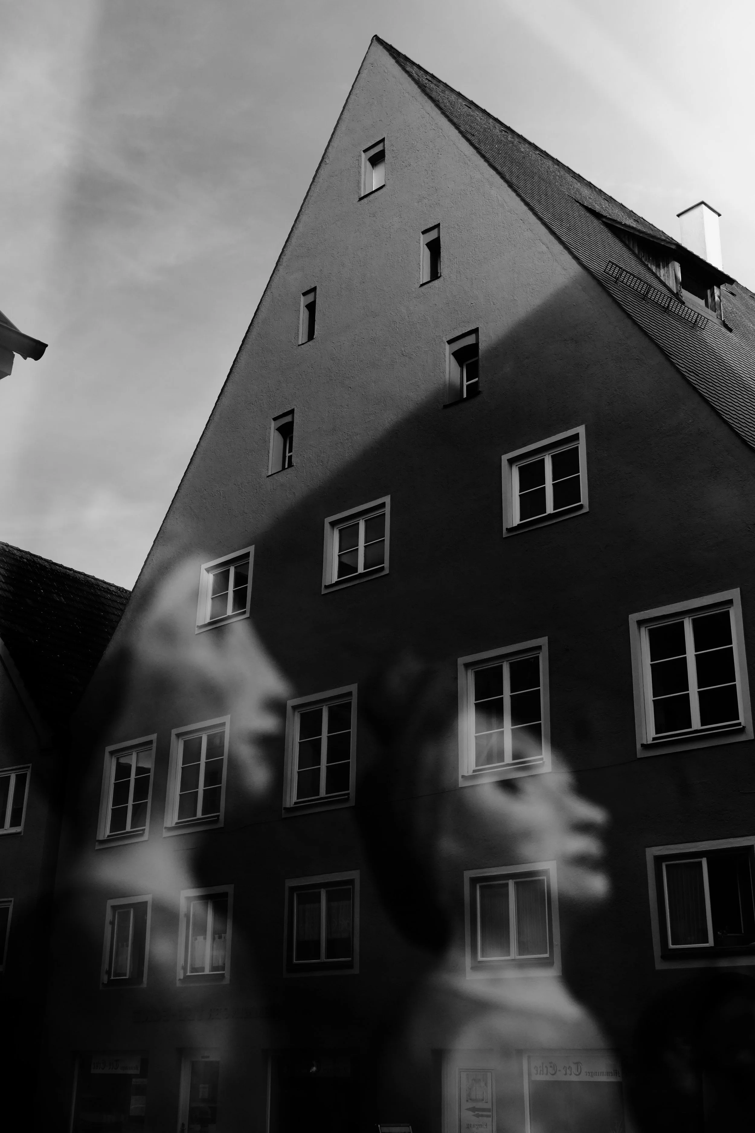 Schwarz-Weiß-Fotografie eines Gebäudes mit schräg stehendem Dach und mehreren Fenstern. Das Bild enthält eine Doppelbelichtung, die eine menschliche Silhouette und Wolken simuliert.