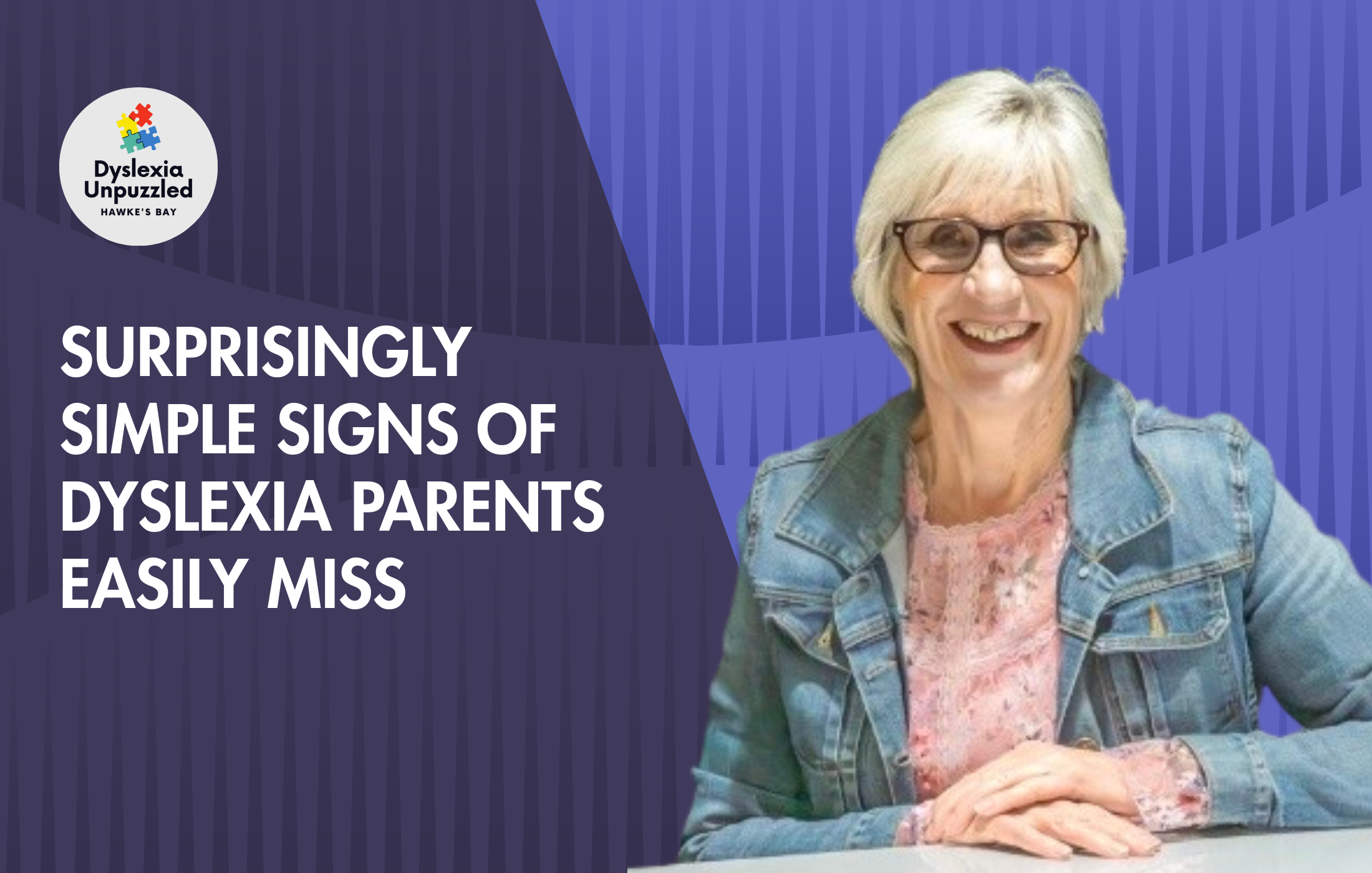 A smiling older woman with short gray hair and glasses, wearing a denim jacket, seated at a table with her hands clasped. Part of a logo with colorful puzzle pieces and text about dyslexia awareness is visible on a purple and blue background.